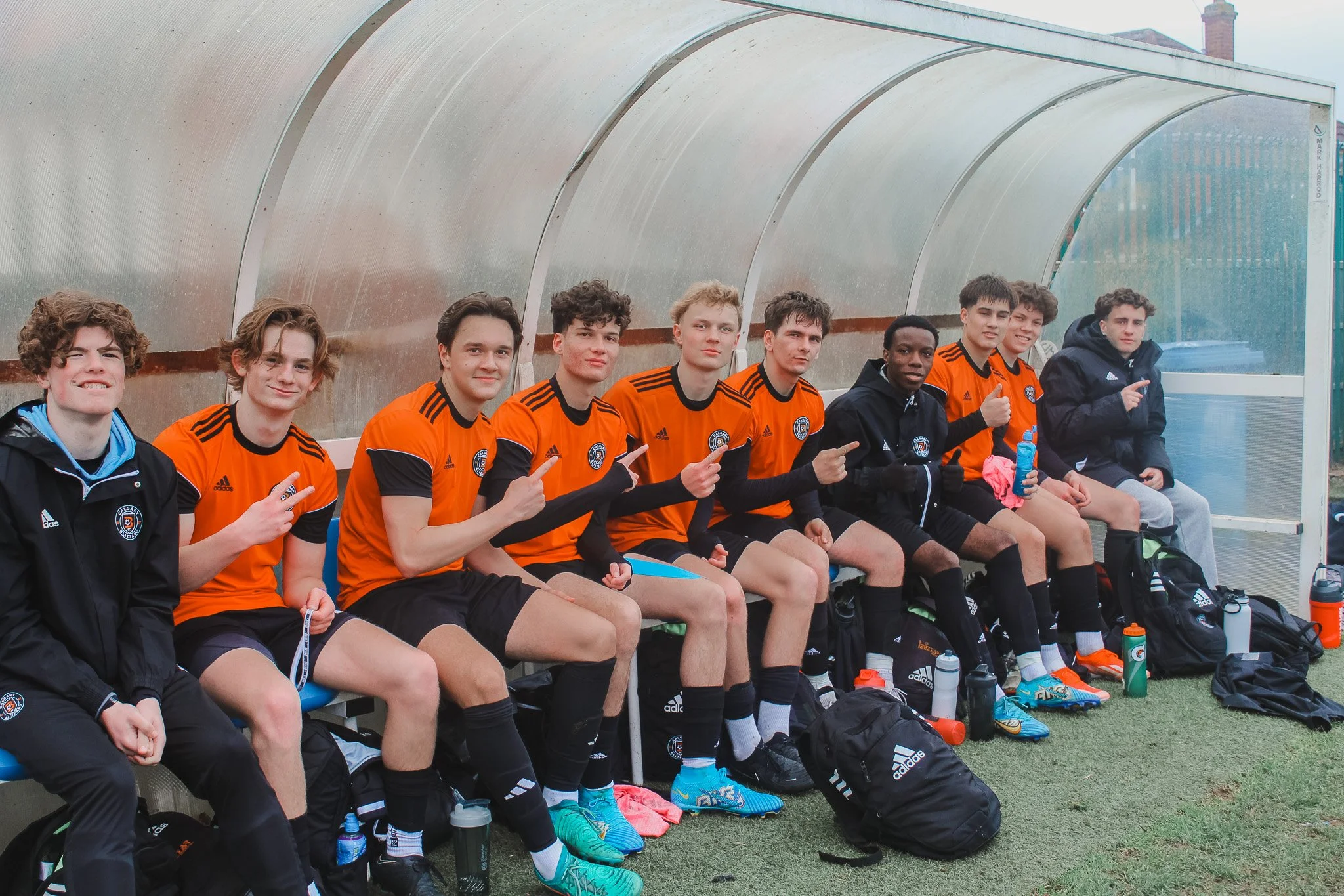 A group of young male soccer players sitting on a bench inside a team dugout, wearing orange and black uniforms with some in black warm-up jackets, with water bottles and gear on the ground in front of them.