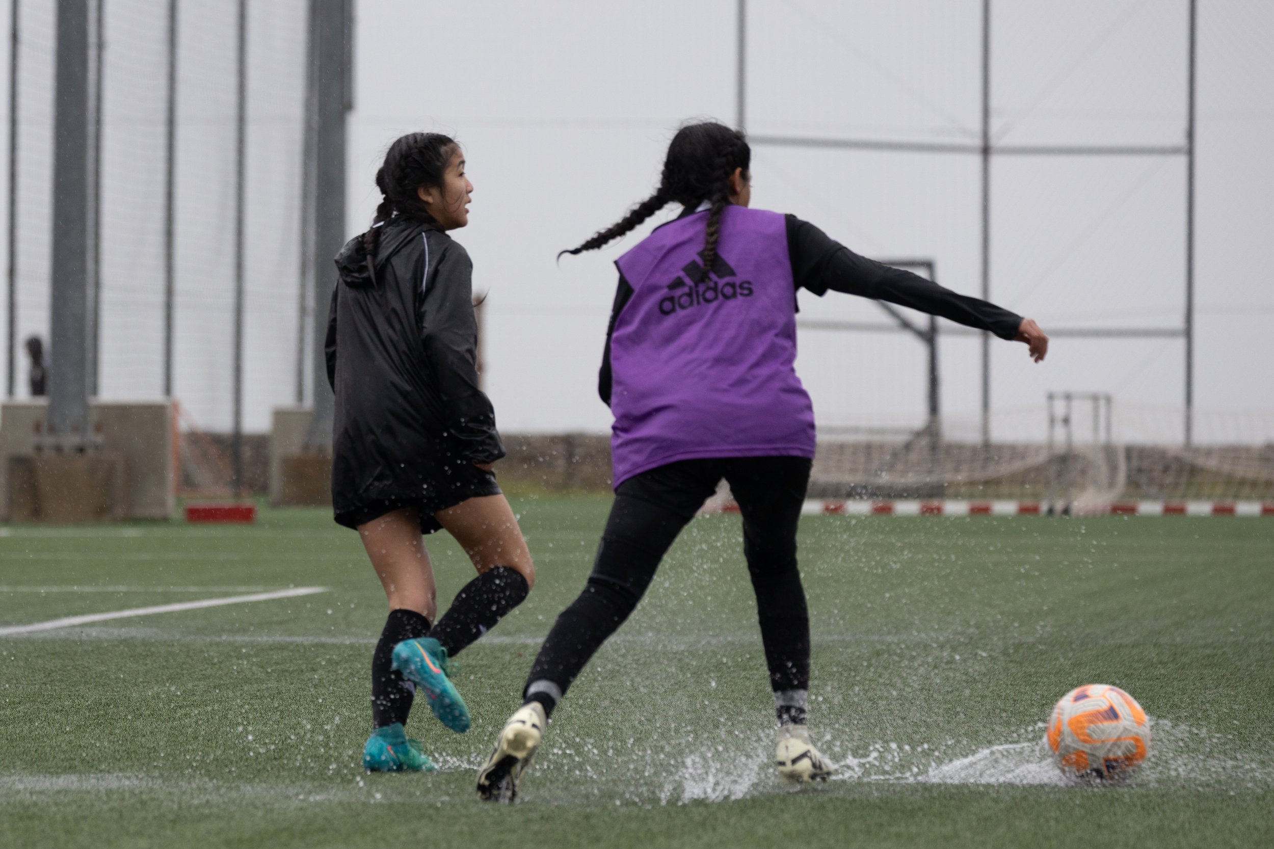 Two young girls playing soccer on a rainy day, one wearing a purple Adidas vest and the other in a black jacket, chasing a soccer ball on a wet field.