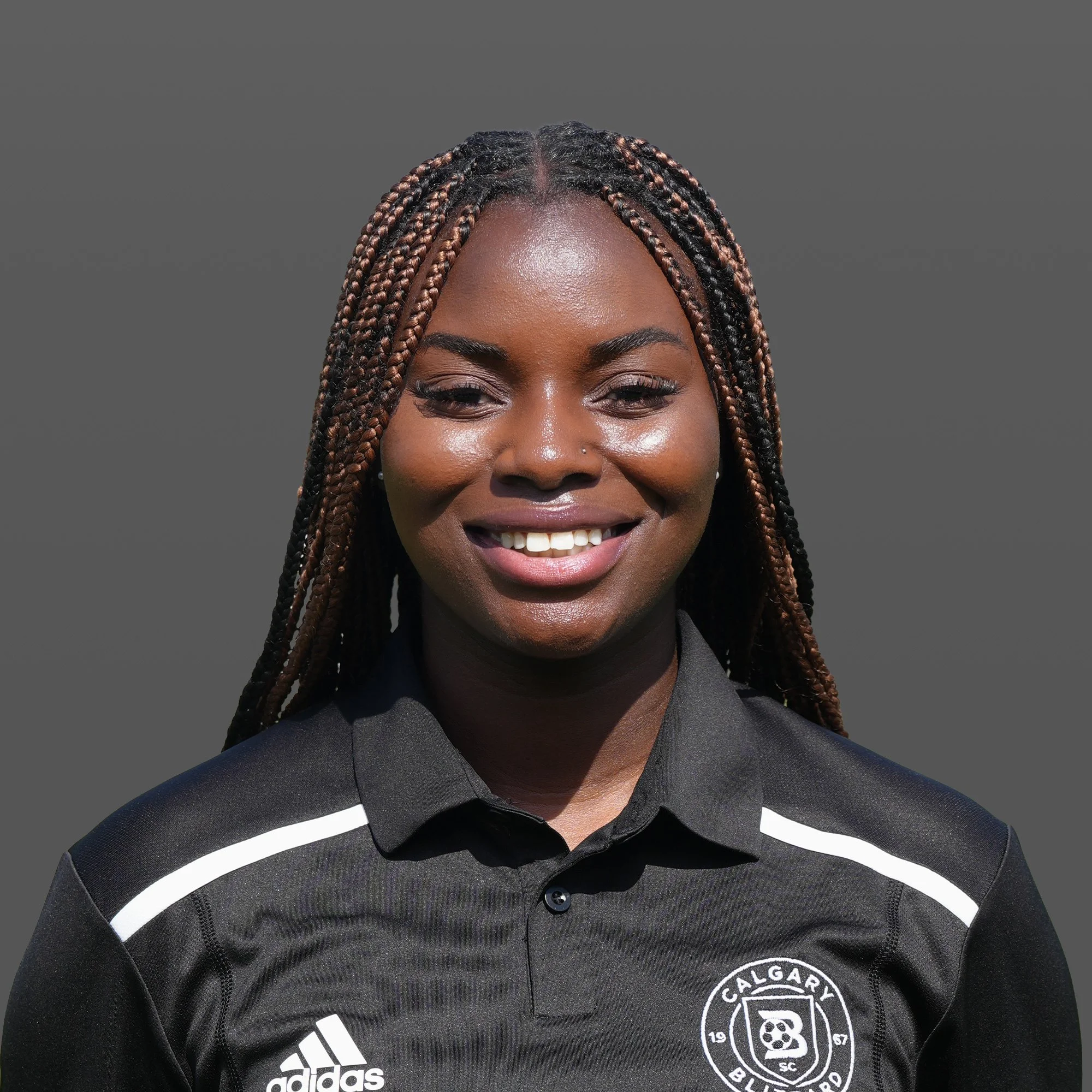 Close-up of a young woman with braided hair, wearing a black sports jacket with a logo and standing on a soccer field at night.