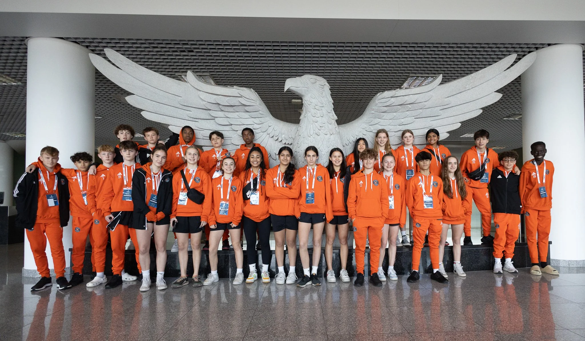 A group of young athletes wearing orange and black sportswear, standing in front of a large sculpture of a white eagle with outstretched wings inside a modern building.