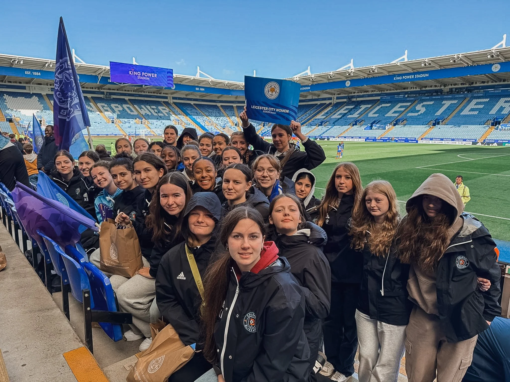 Group of women and girls at a soccer stadium, some holding flags and a sign that reads "Leicester City Women." The stadium seating is mostly empty, and the field is visible in the background.