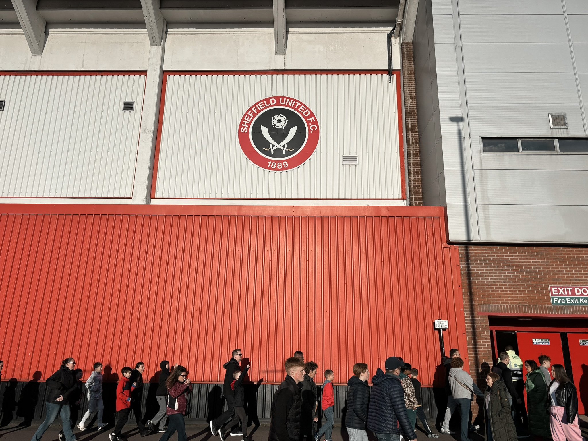 Crowd of people walking past the Sheffield United Football Club stadium with its logo and red exterior.