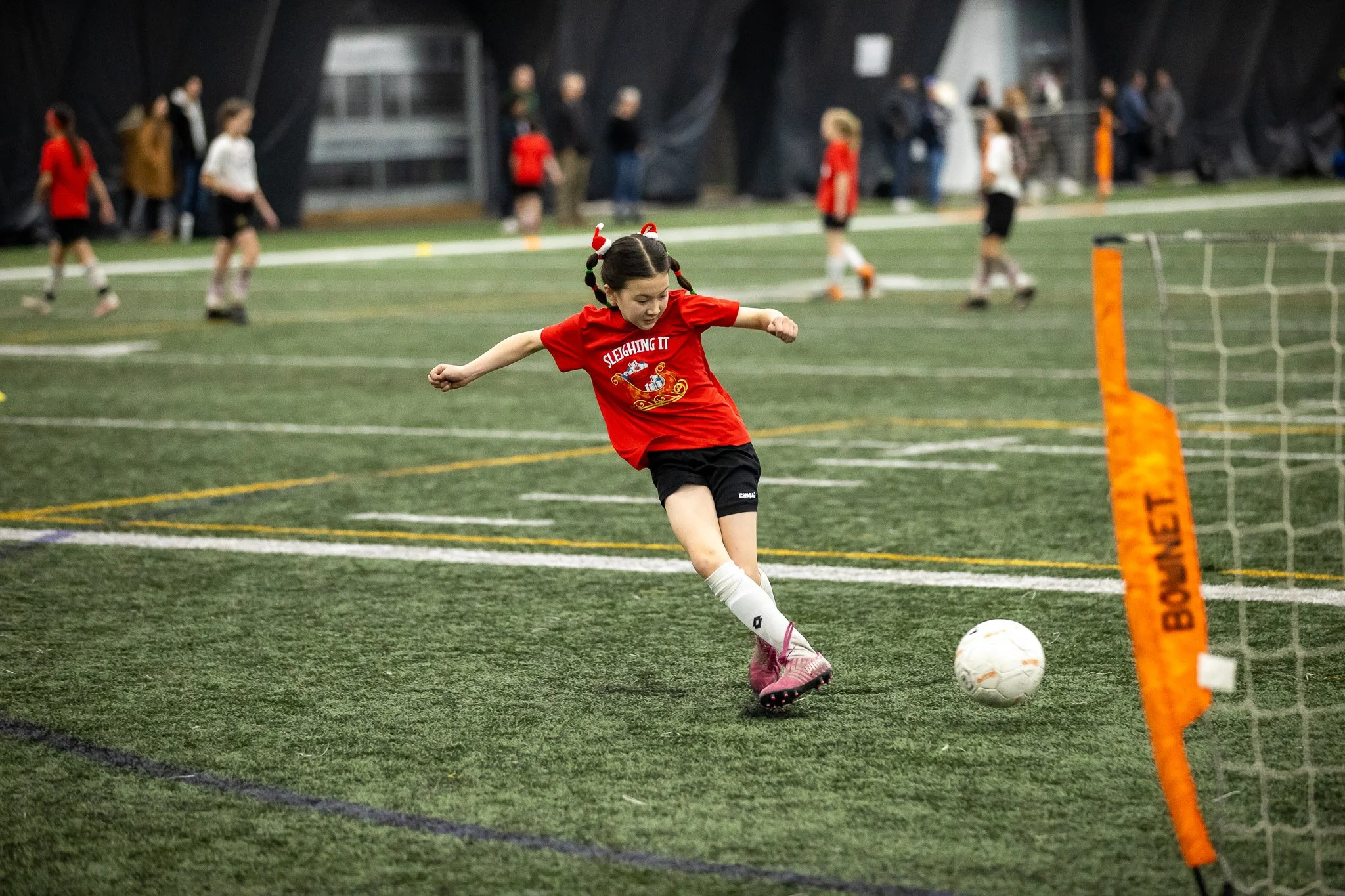 A young girl wearing a red shirt and black shorts is kicking a soccer ball on an indoor synthetic turf field, with several children and adults in the background.