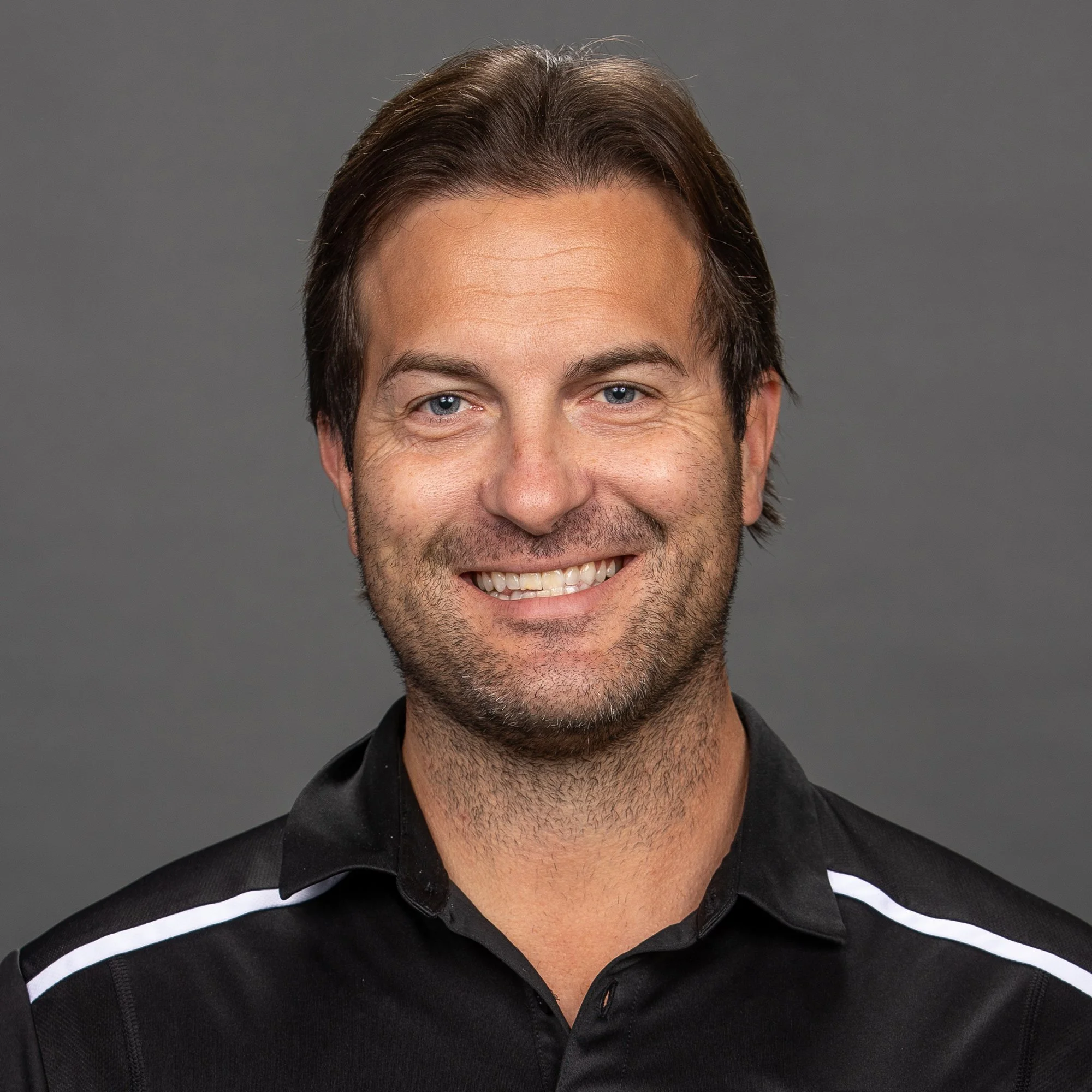 Portrait of a smiling man with brown hair and blue eyes, wearing a black sports jacket with patches, standing outdoors during early evening or night with blurred lights in the background.