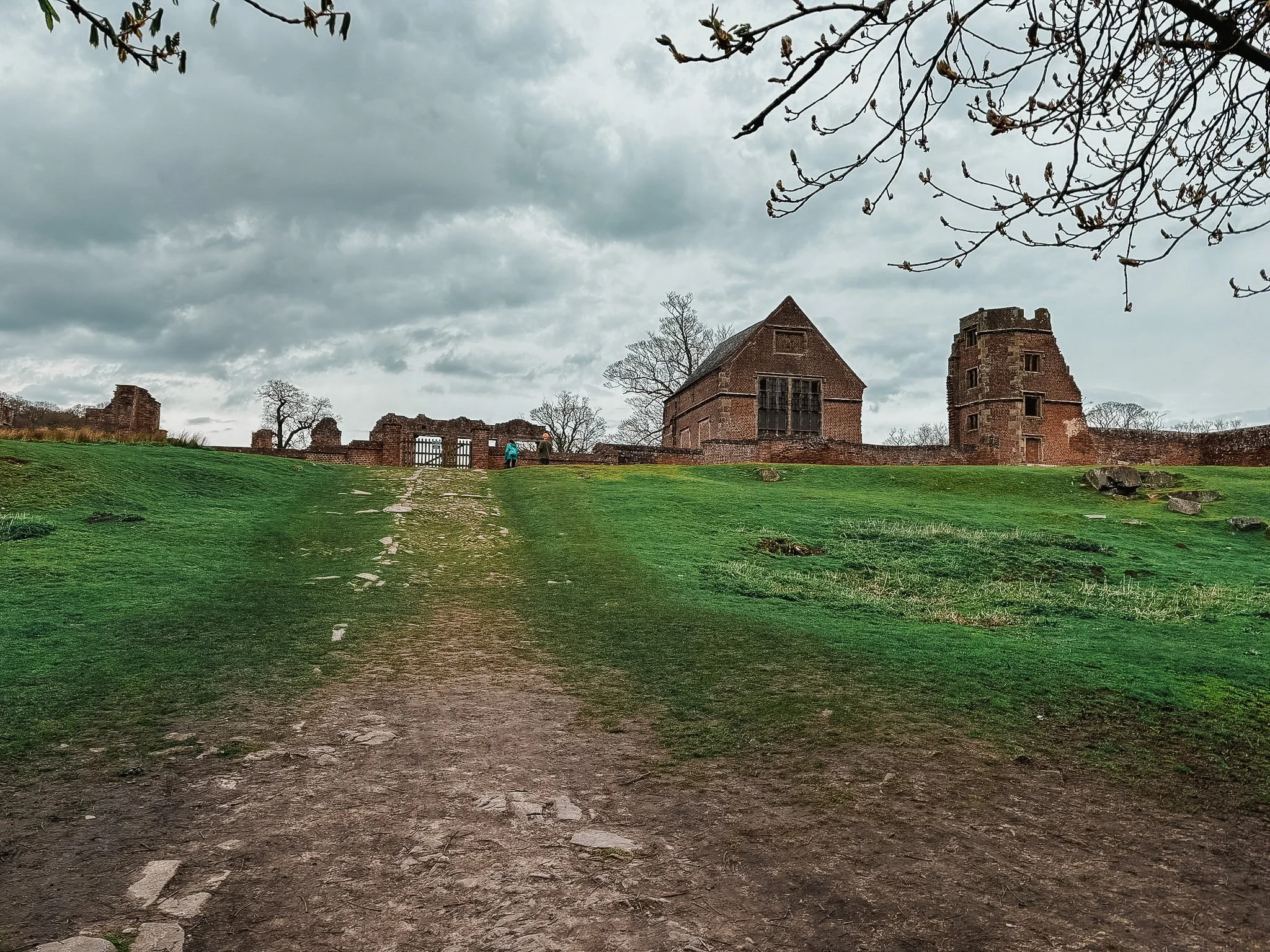 Ancient castle ruins on a grassy hill under a cloudy sky, with a dirt path leading up to the structures.