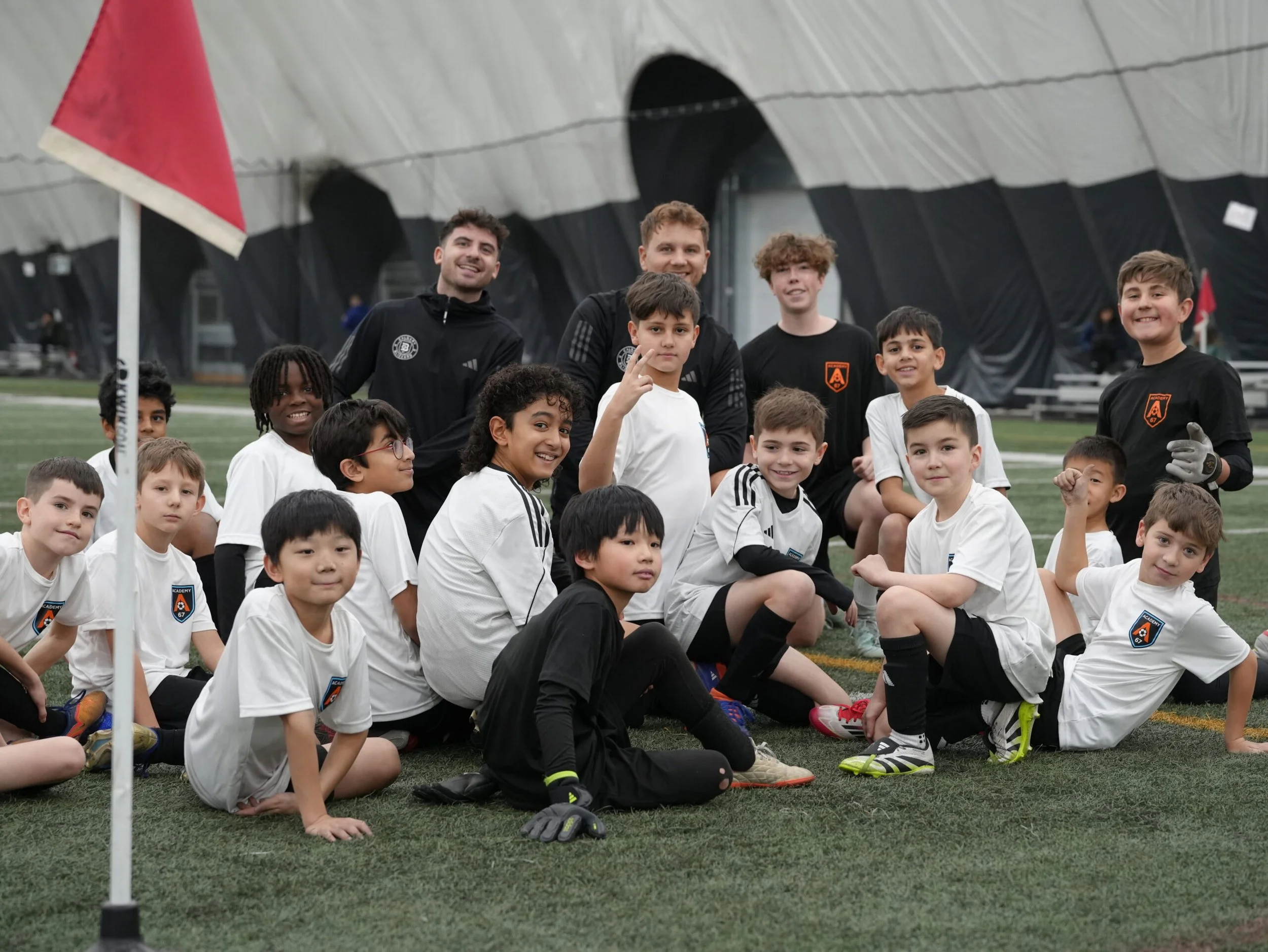 A diverse group of children and adult coaches posing for a team photo on an indoor soccer field, some kneeling, sitting, or standing, in front of a black and orange soccer goal, with the players wearing white and black uniforms.