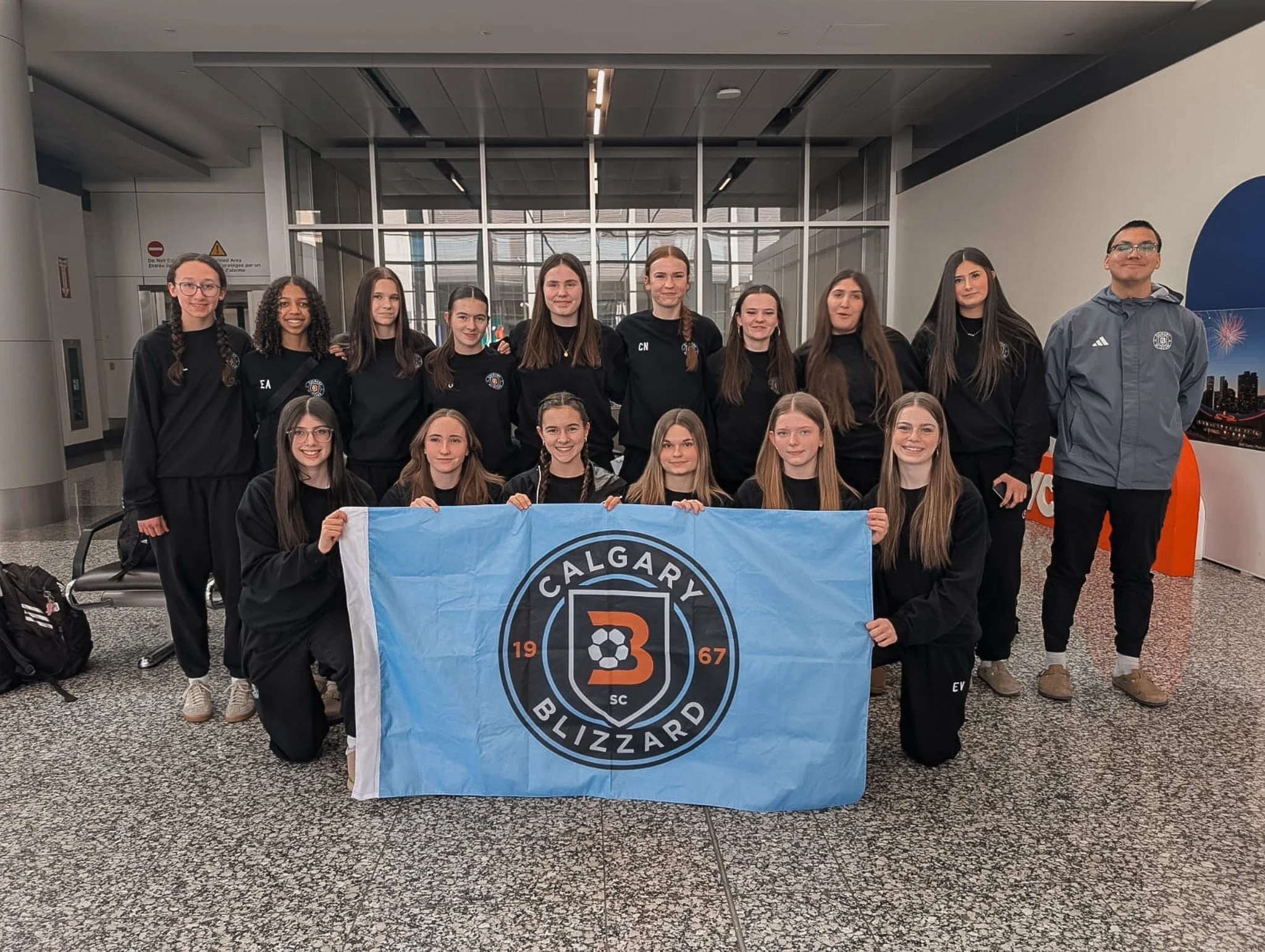 A group of young women and a man holding a Calgary Blizzard soccer team flag at an indoor location, likely an airport or station.