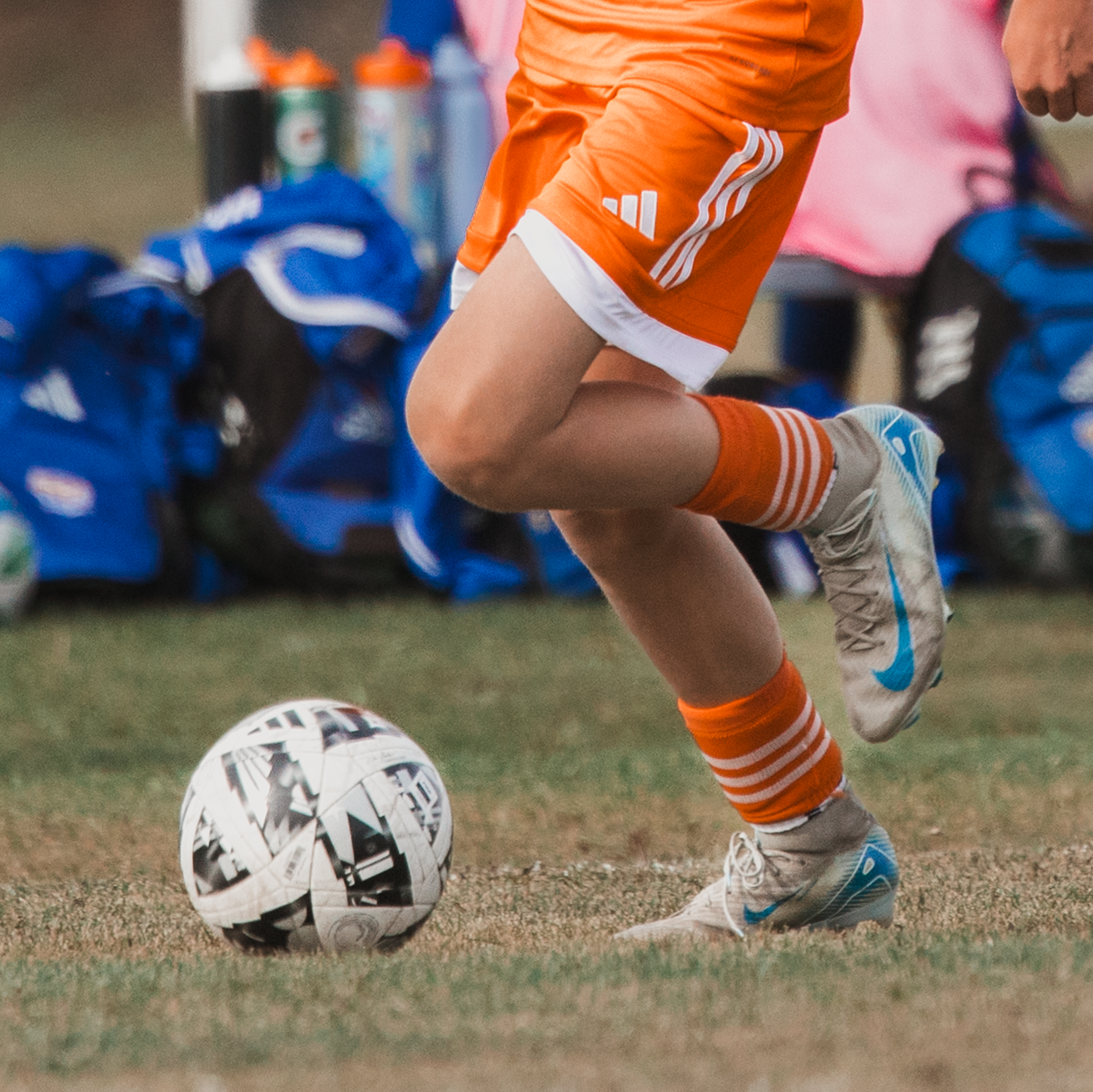 Close-up of a soccer player's legs, dressed in orange and white uniform with Nike shoes, kicking a black and white patterned soccer ball on a grass field with blue bags and water bottles in the background.