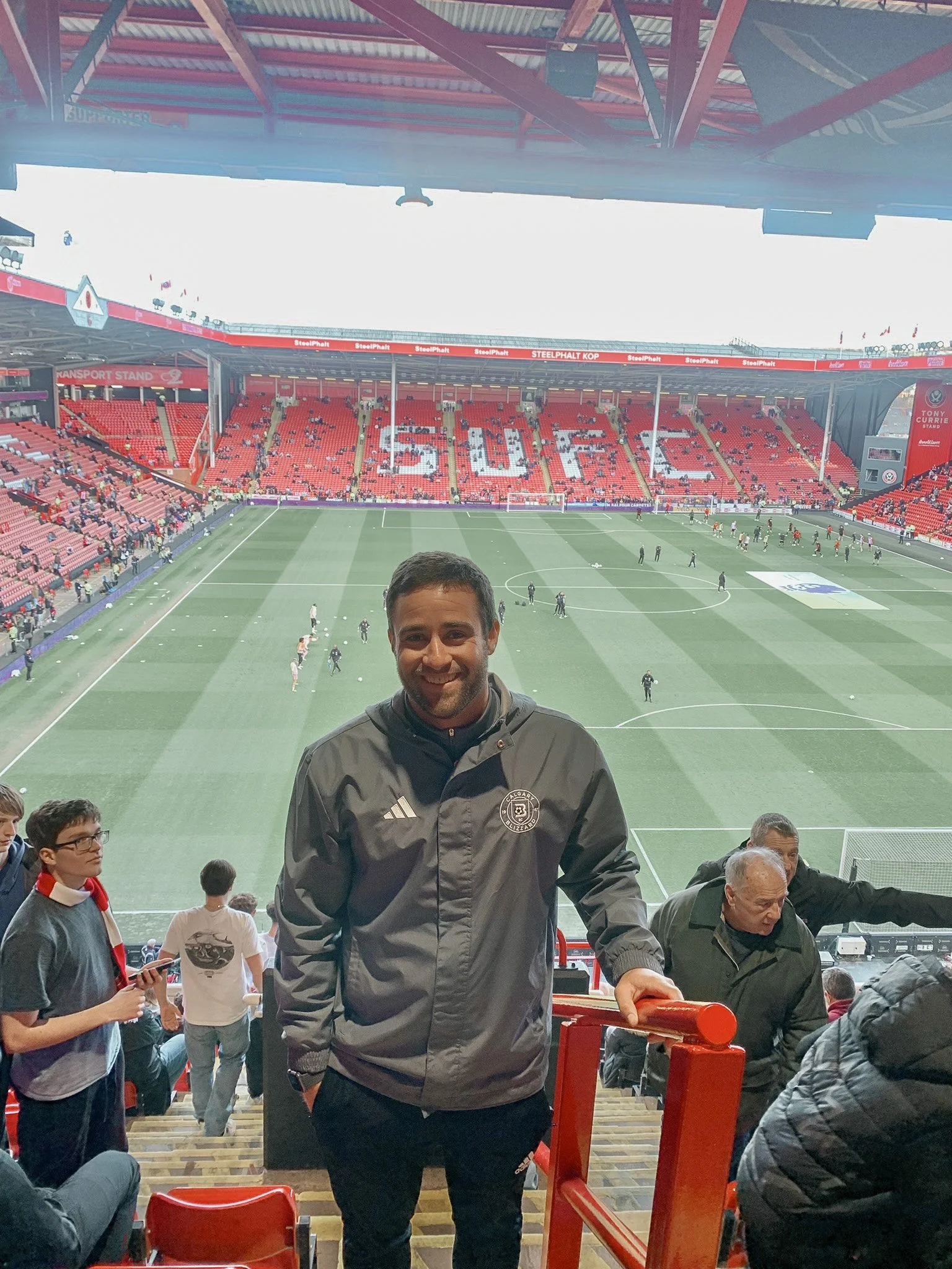 A man smiling at the camera standing in a stadium with a soccer field in the background, spectators around him, and the word 'CELTIC' spelled out in the stands.