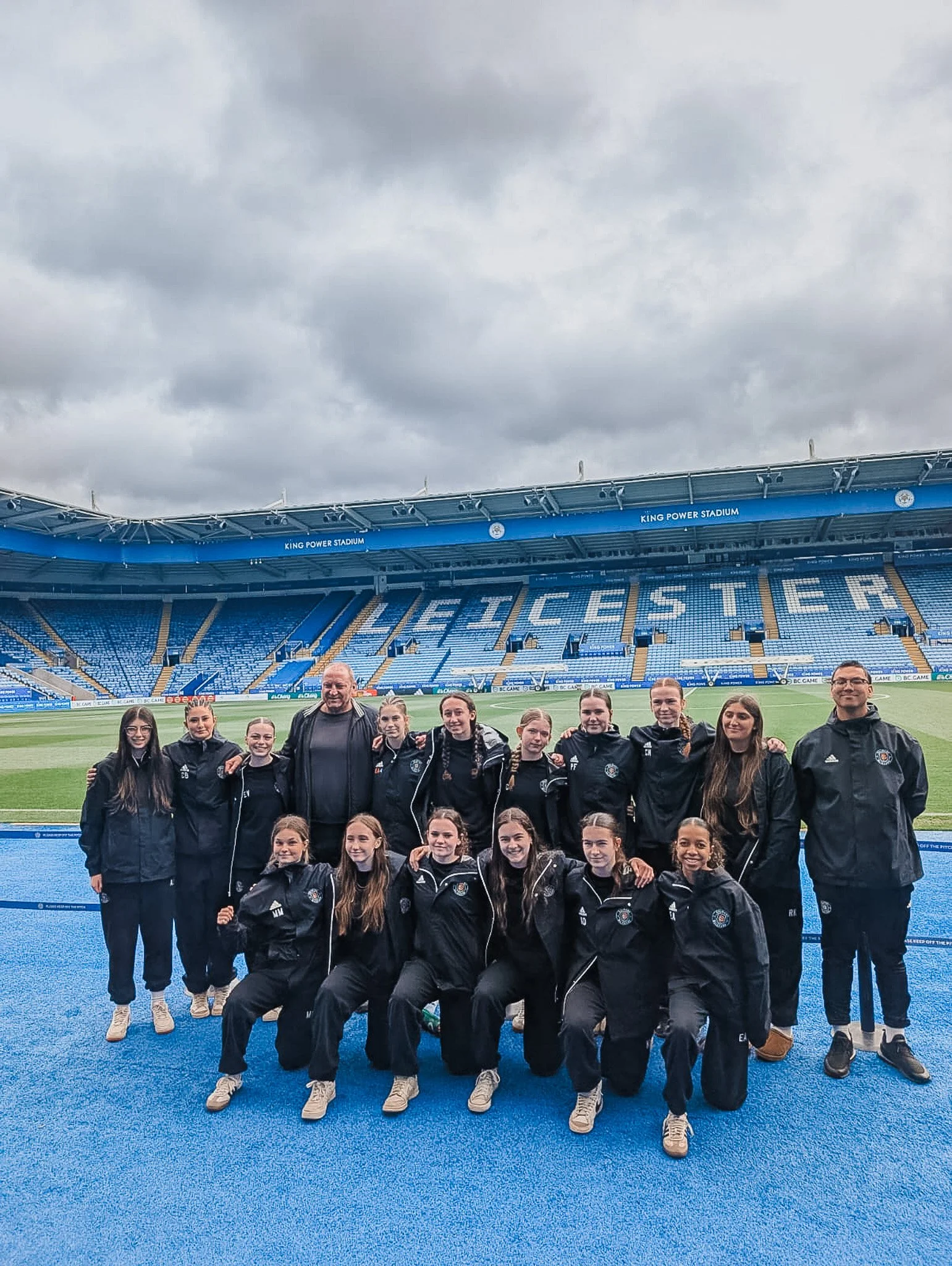 Group of young female athletes and coaches in matching black jackets, posing on a blue field at King Power Stadium in Leicester, under a cloudy sky.