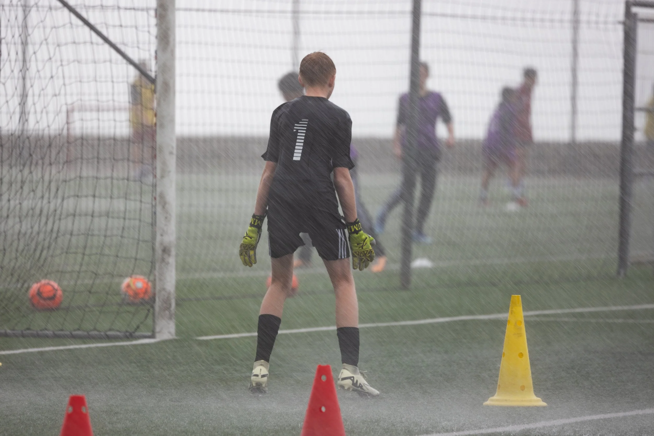 A young soccer goalkeeper in black uniform with the number one on the back, wearing gloves, standing in front of the goal during a training session in the rain. Two orange soccer balls are in the goal, and colored cones are on the ground for drills. Other players are seen in the background on the field.