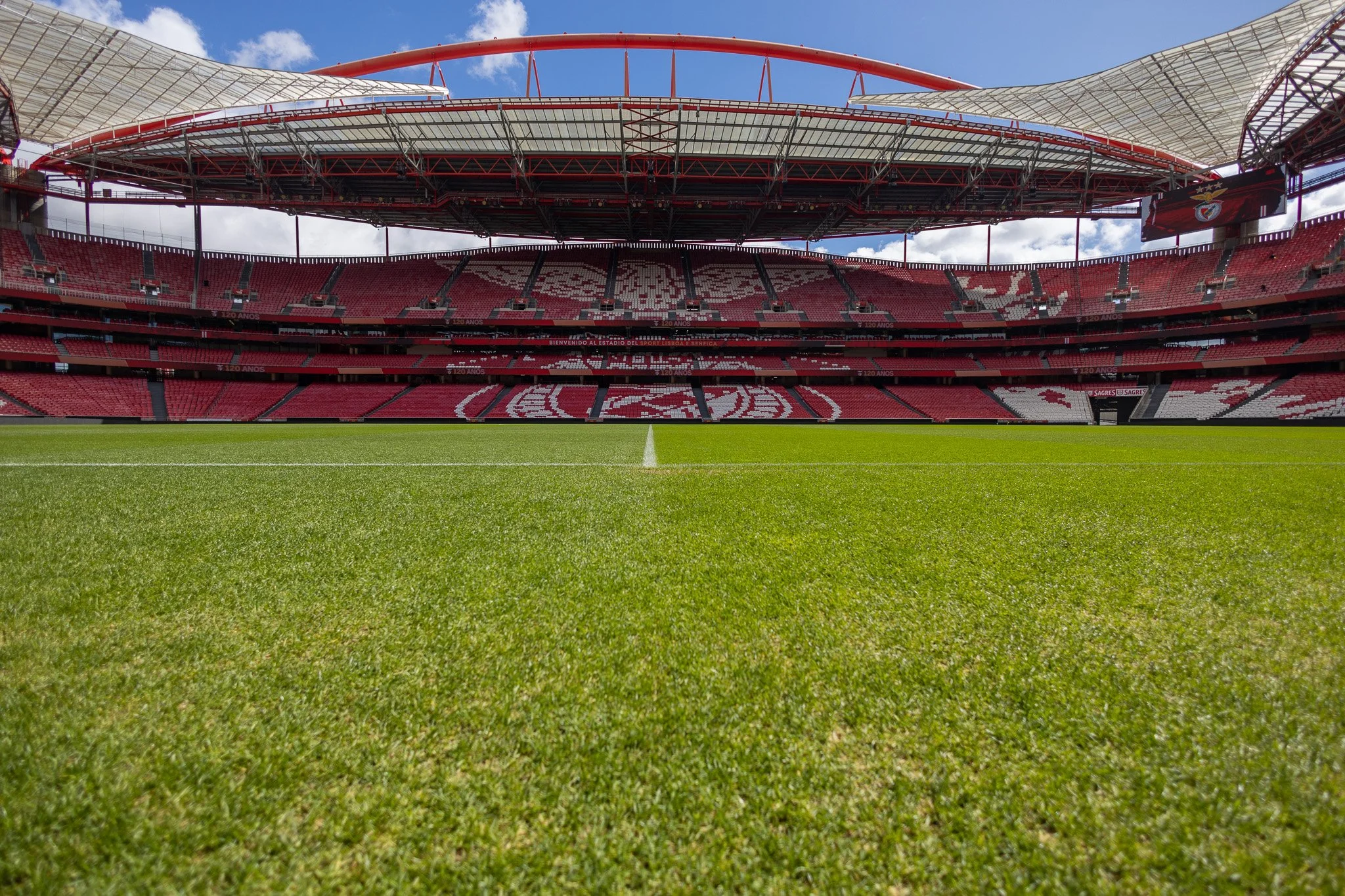 Empty soccer stadium with green field and red seats, with large, partially covered roof and cloudy sky.