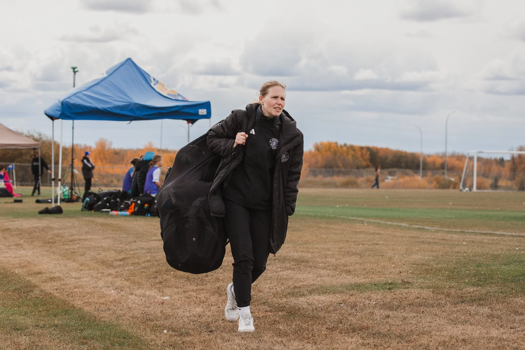 A woman carrying a large black sports bag walking on a grassy field with a blue tent, other people, and trees with autumn foliage in the background.