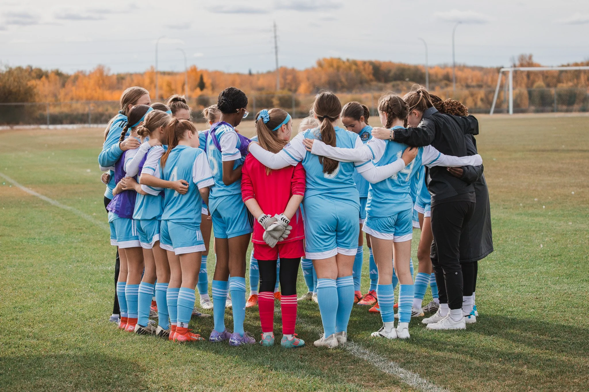 A girls' soccer team huddles together on the field during daytime, with some players wrapping their arms around each other, listening to their coach.
