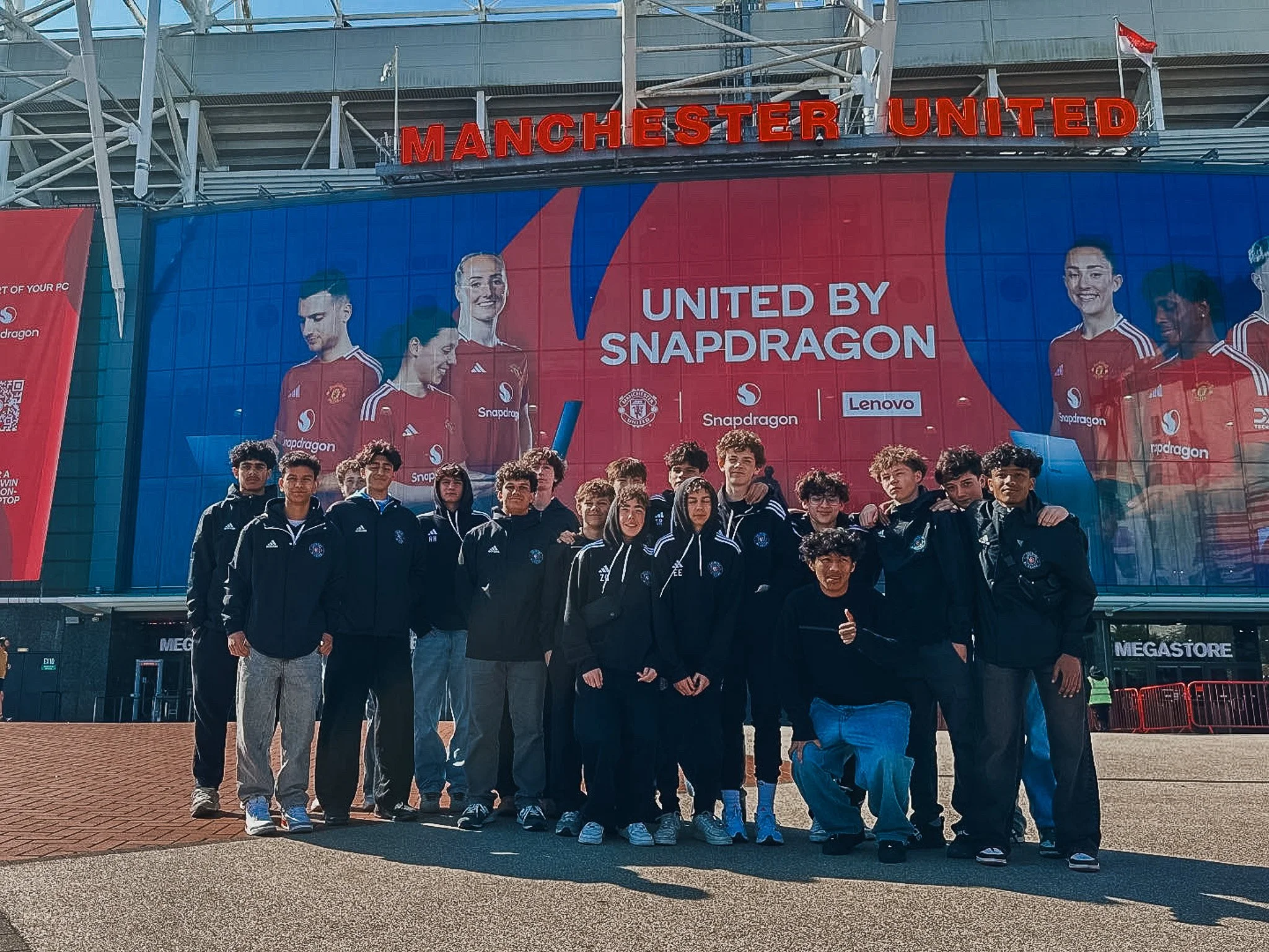 Group of young people posing in front of Manchester United stadium with a large banner displaying players and the text "United by Snapdragon".