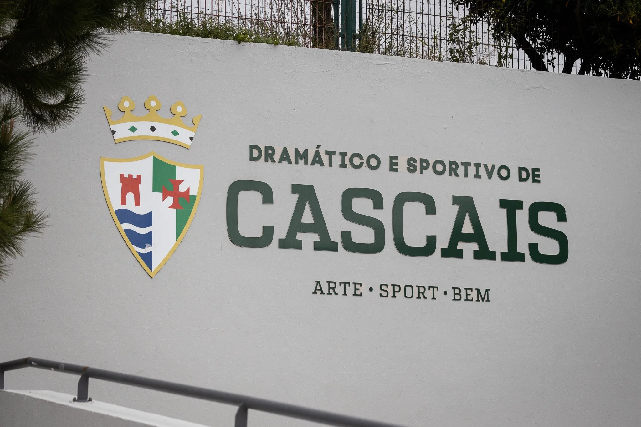 Photograph of a white wall with a crest featuring a crown, a red castle, a green and white banner, and blue waves, along with the text "Dramático e Sportivo de Cascais" and the words "Arte, Sport, Bem" beneath.