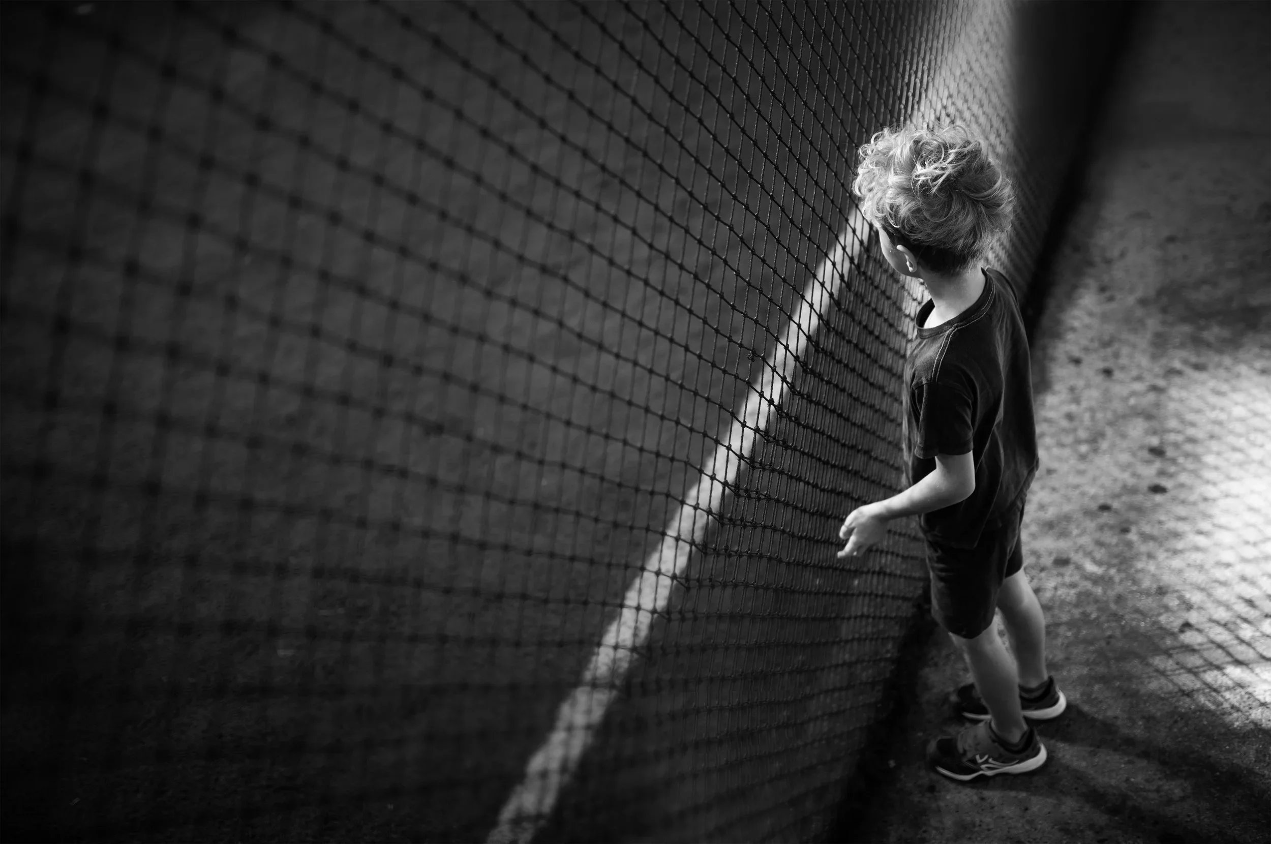 A boy with curly hair standing next to a tennis net, looking over the net on an outdoor tennis court.