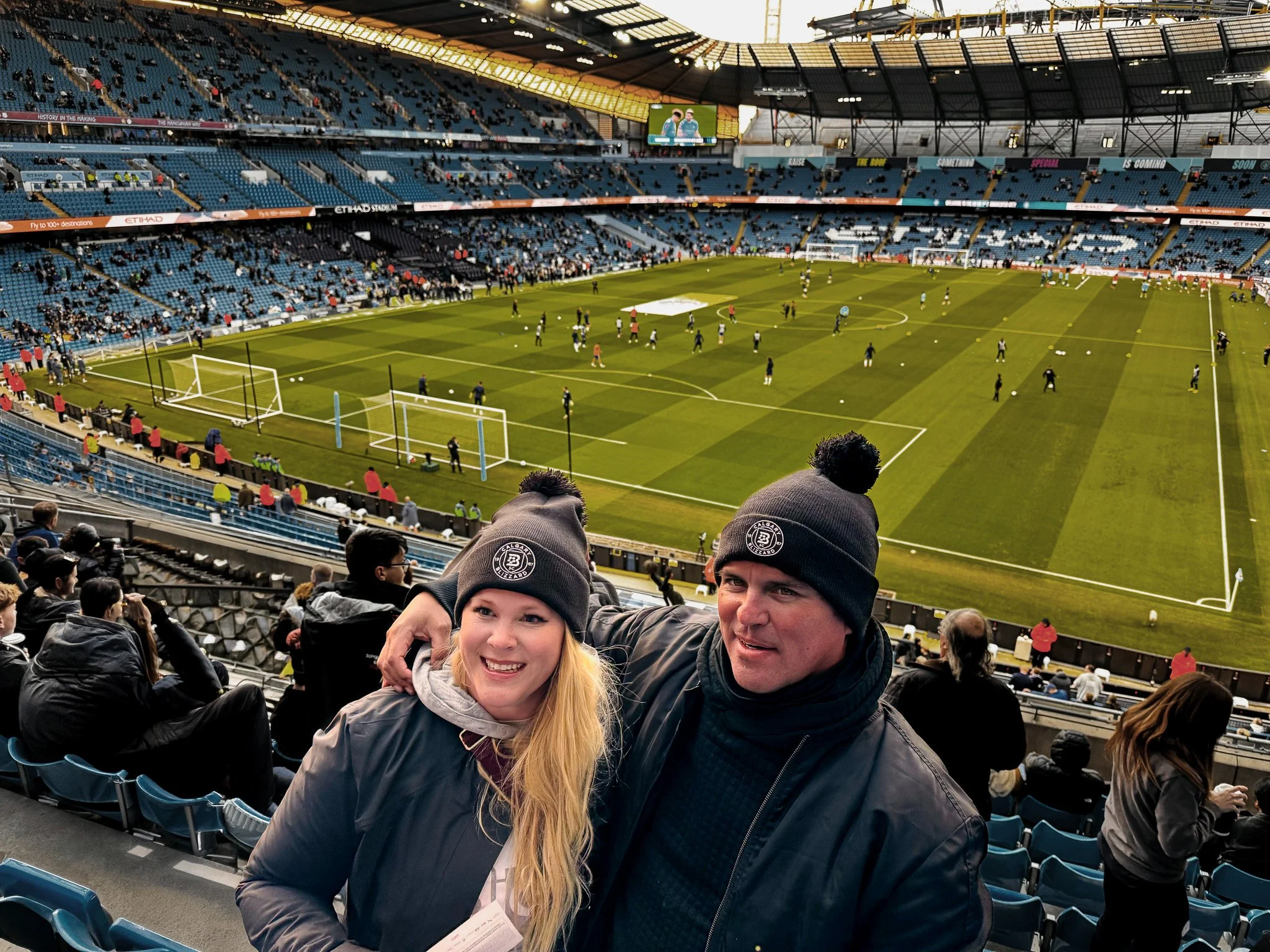 A smiling woman and man wearing black beanies with a team logo, standing in a stadium with a soccer field in the background. The stadium has spectators and players warming up on the field.