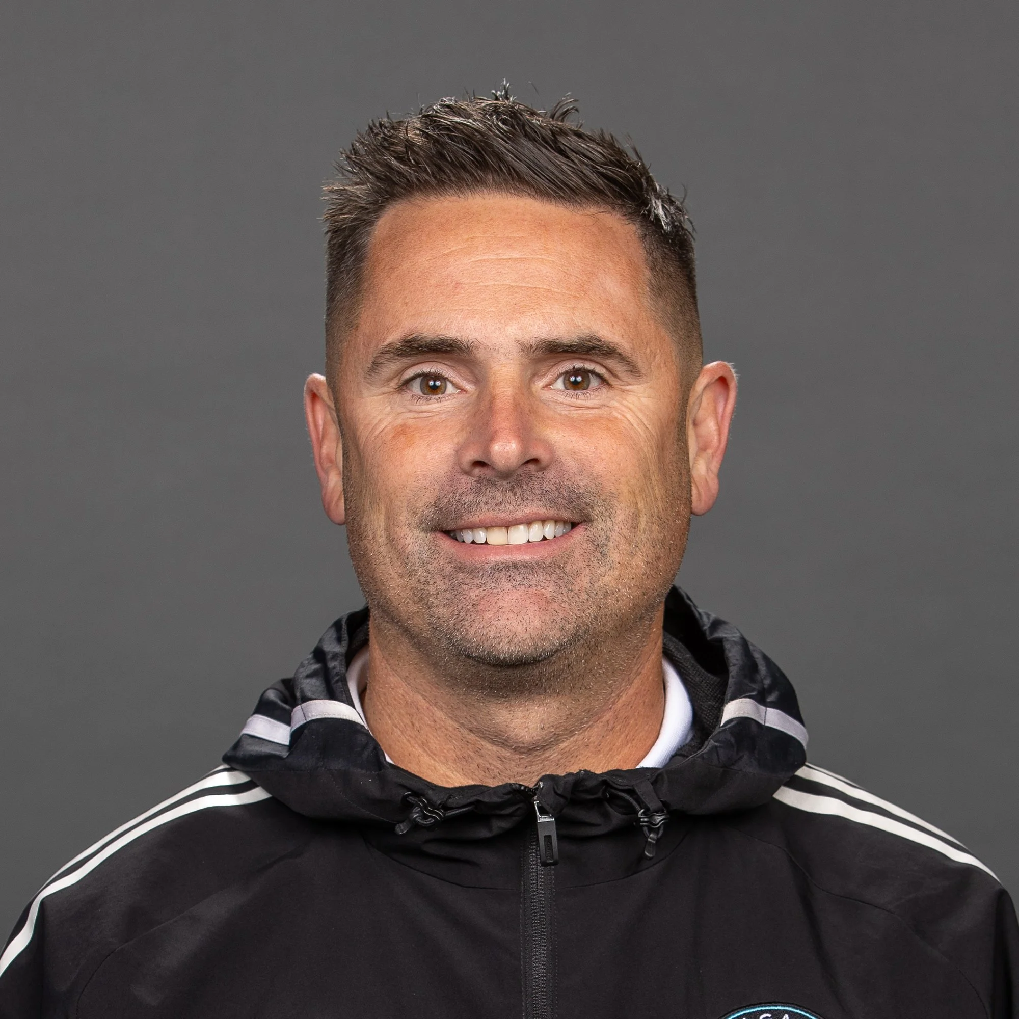 A man with short dark hair and blue eyes wearing a black Adidas sports jacket with a badge on the chest is standing outdoors at night.