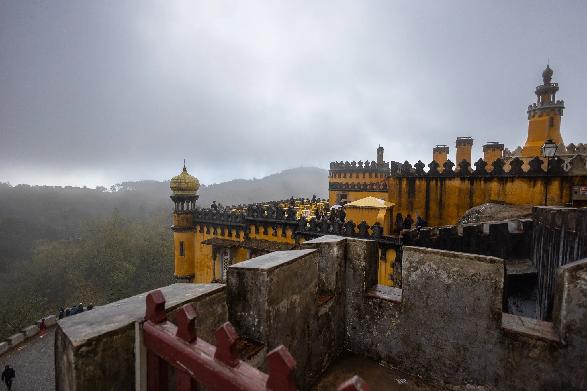 Historic yellow castle with dark accents on a foggy hillside, featuring turrets and battlements, with visitors on the walkway.