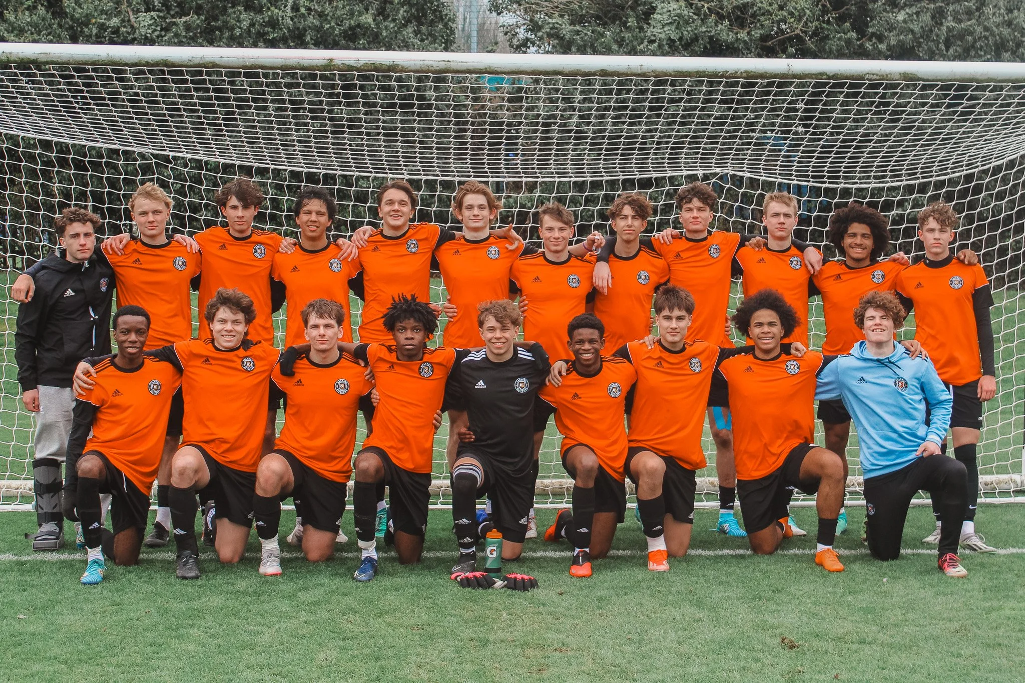 A youth soccer team poses for a photo in front of a goal on a grassy field, wearing orange jerseys and black shorts, with a coach or teammate in a blue jacket kneeling in front.