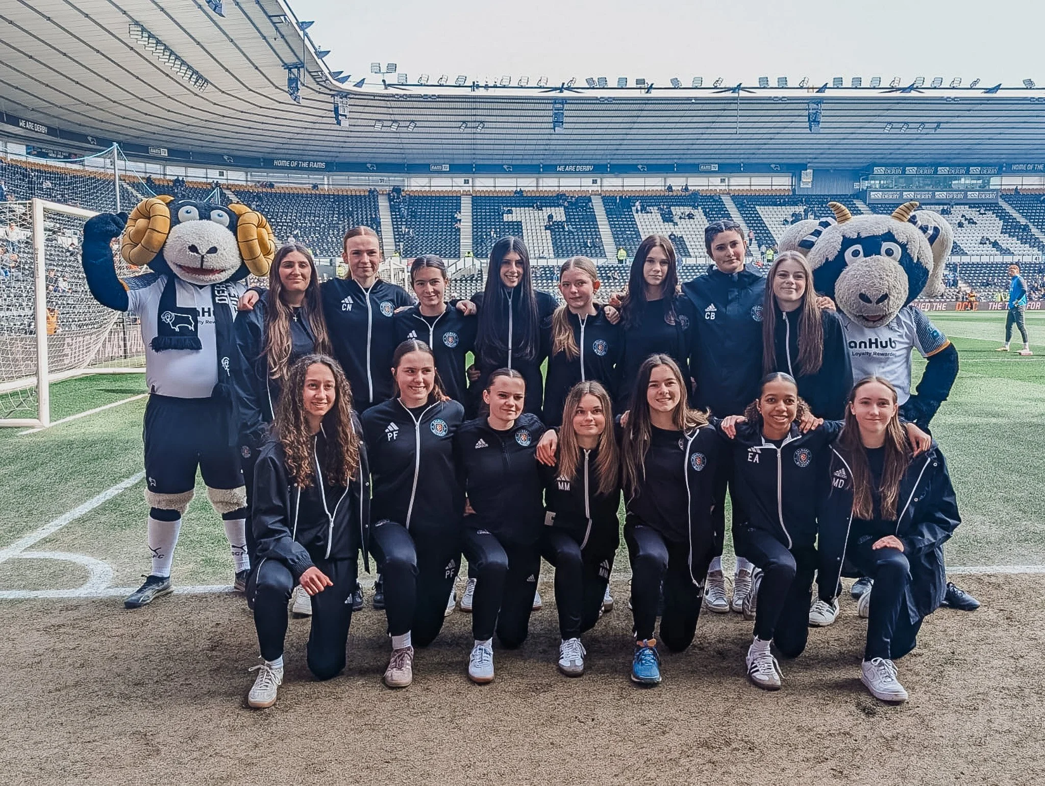 A group of young female soccer players in navy tracksuits posing on a soccer field with two mascots in lion costumes, in a stadium with empty blue and white seats.