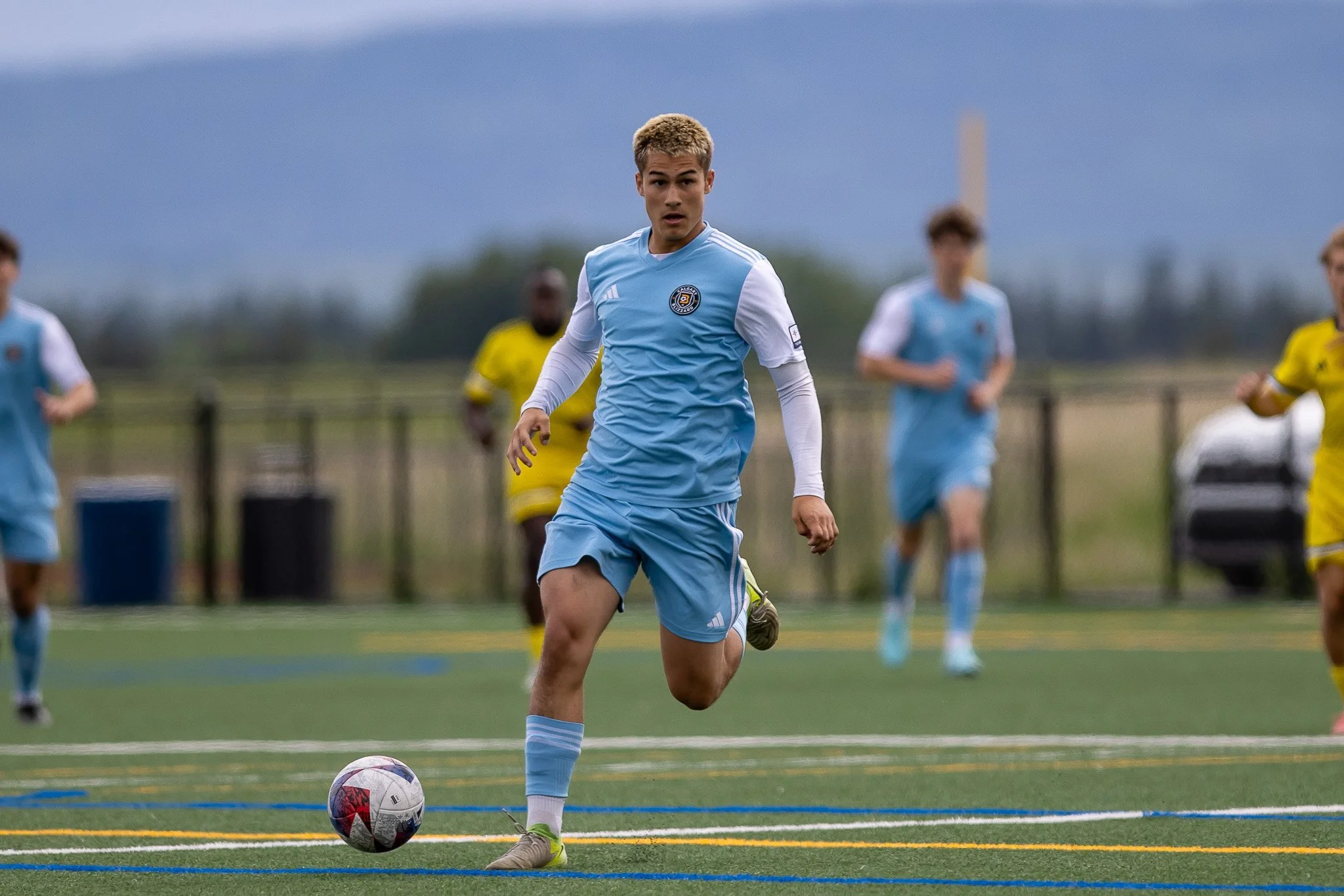 A soccer player in a light blue uniform kicking a ball during a game on an outdoor field, with other players and a fence in the background.