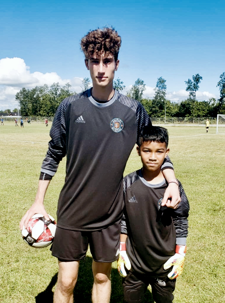 Two boys in black soccer uniforms standing on a grassy field, with one holding a soccer ball. They are posing with their arms around each other, under a blue sky with some clouds, with goalposts and other children playing soccer in the background.