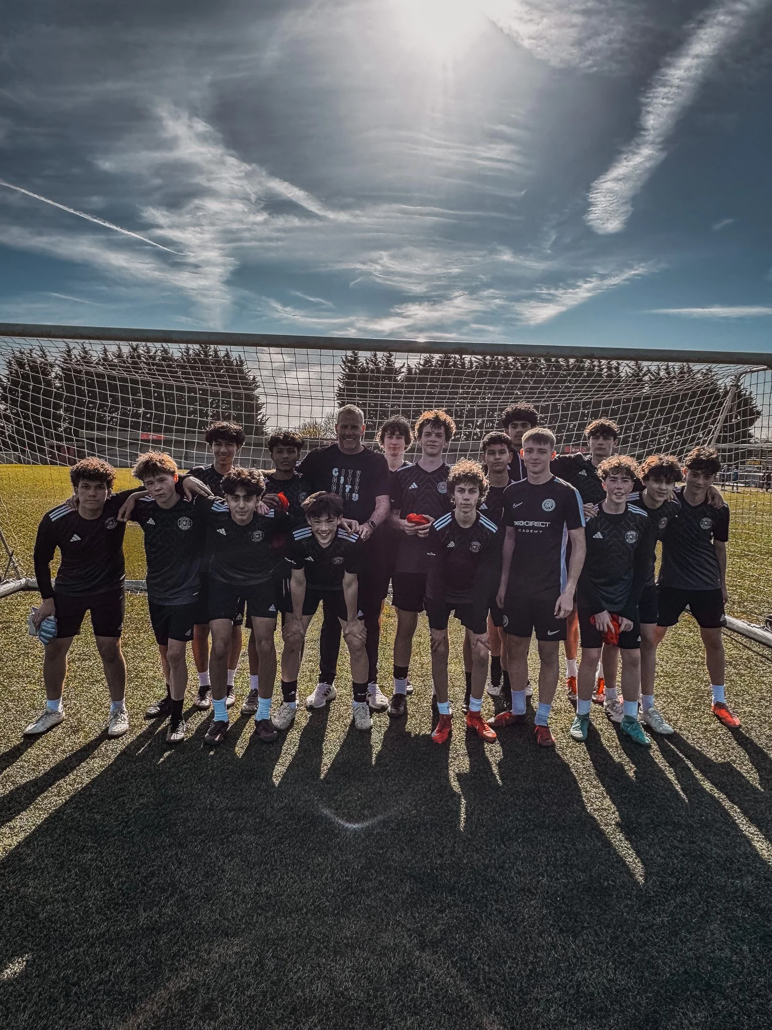 A group of teenage boys and one adult in black soccer uniforms and shorts standing together on a soccer field in front of a goalpost, during daytime with a partly cloudy sky.