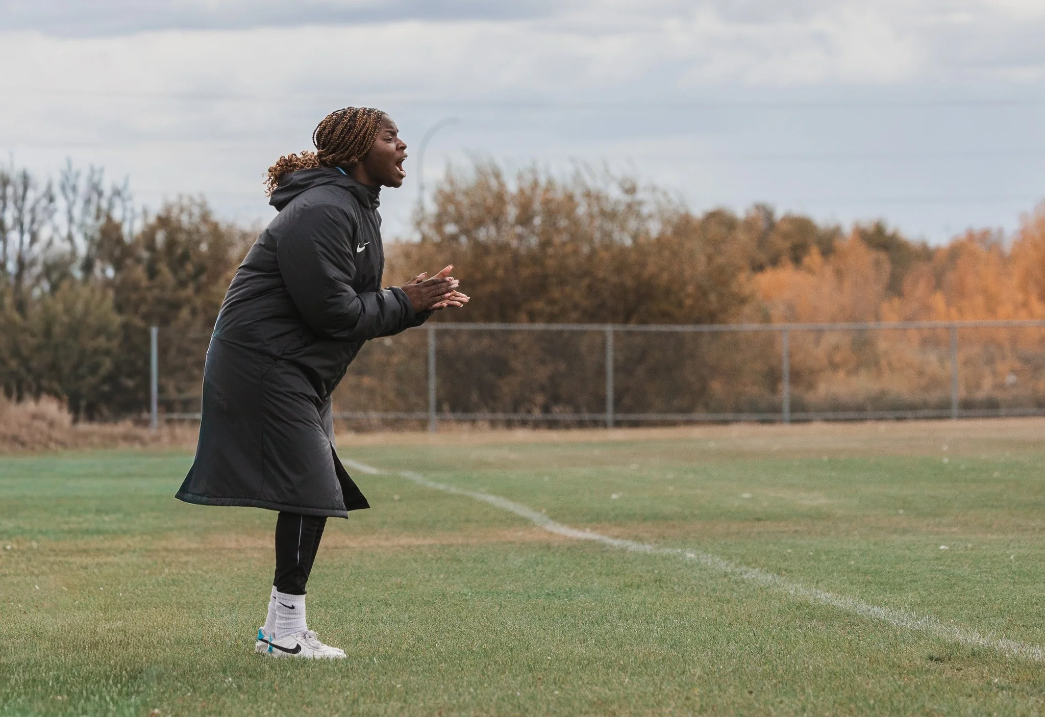 A woman in a black jacket and white sneakers is standing on a soccer field, shouting or coaching.