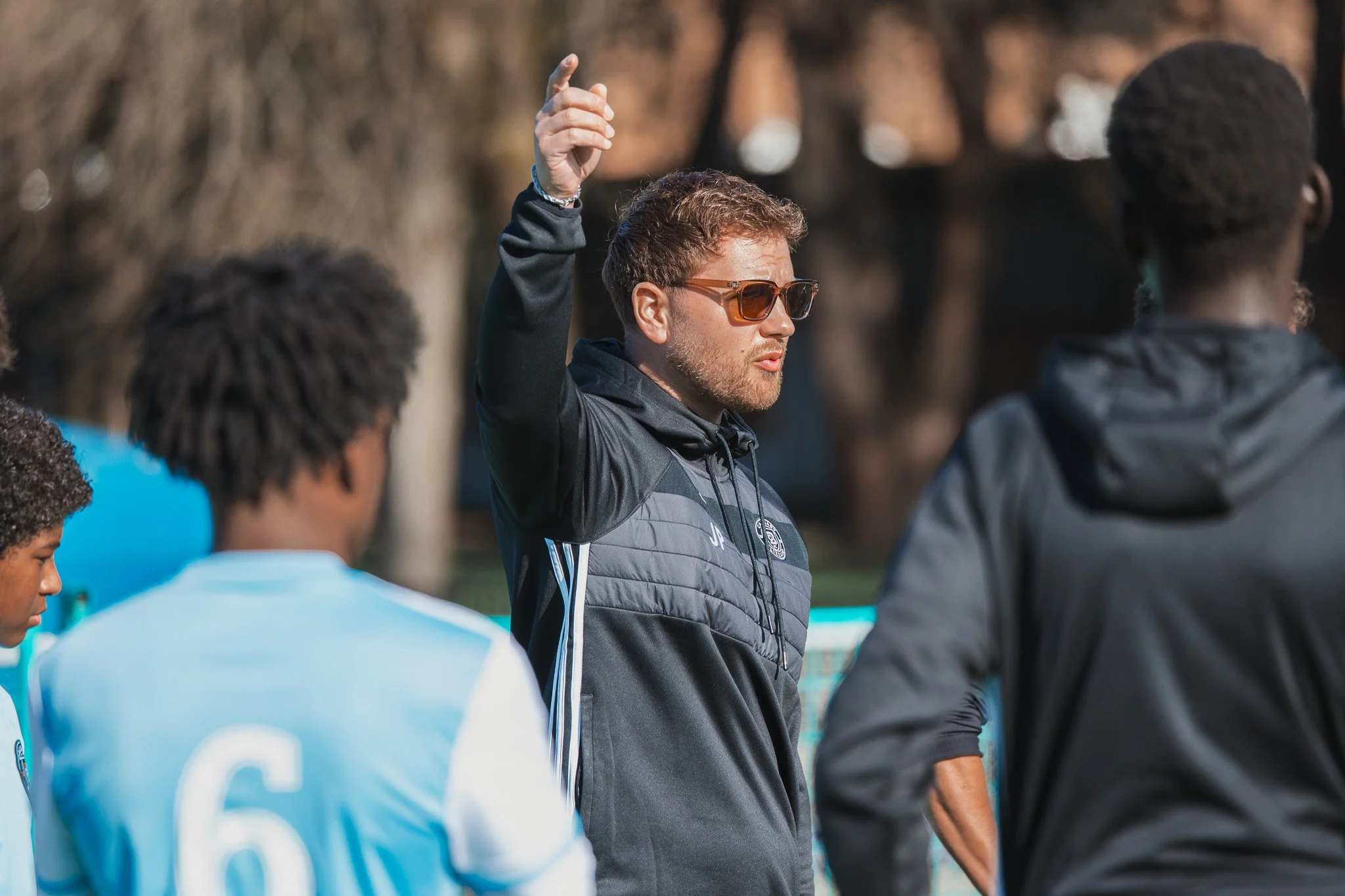A man with sunglasses giving instructions or coaching a sports team on a field, with players in light blue uniforms around him.