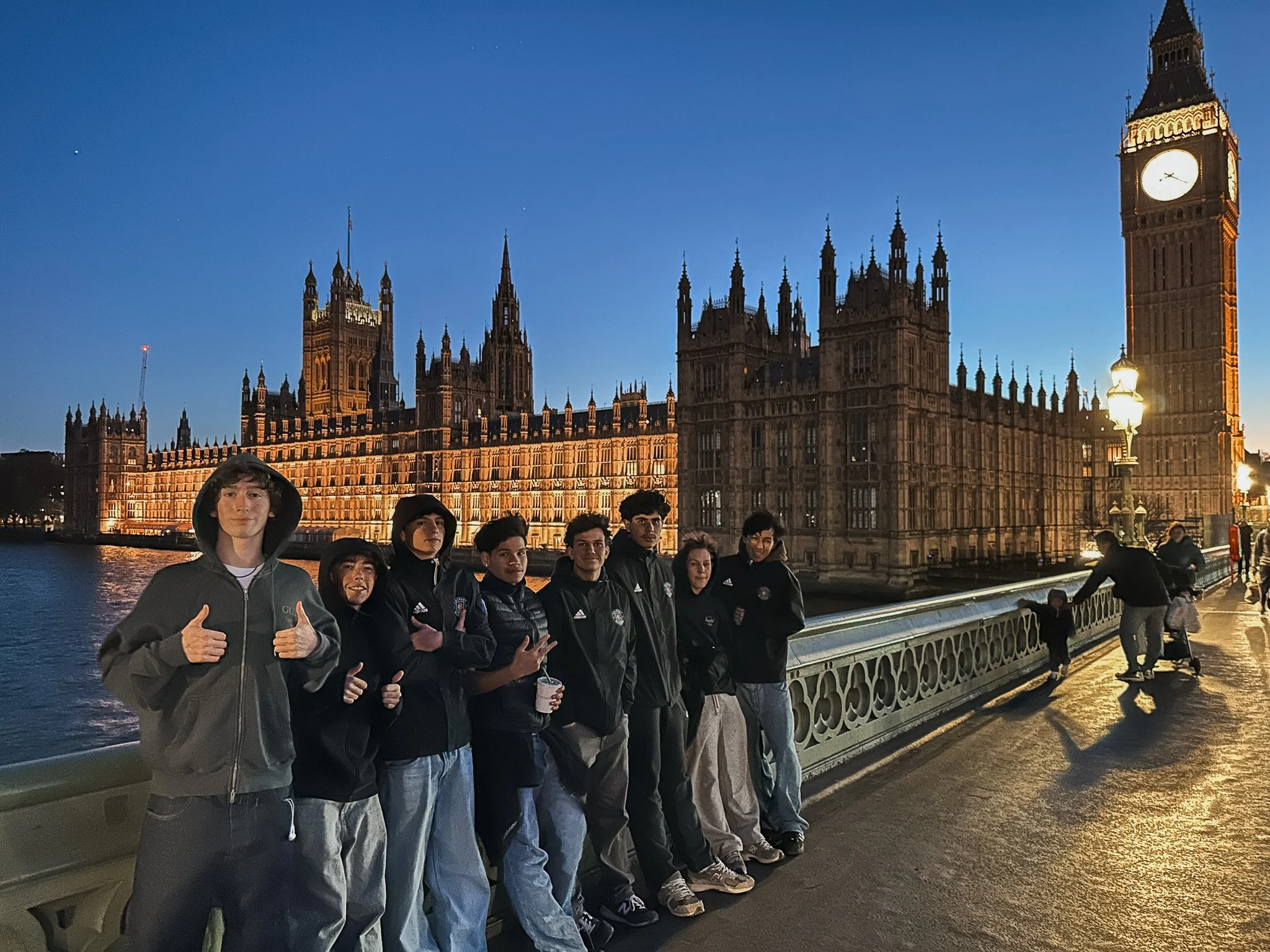 Group of young people standing on Westminster Bridge in London at dusk with the illuminated Palace of Westminster and Big Ben in the background.