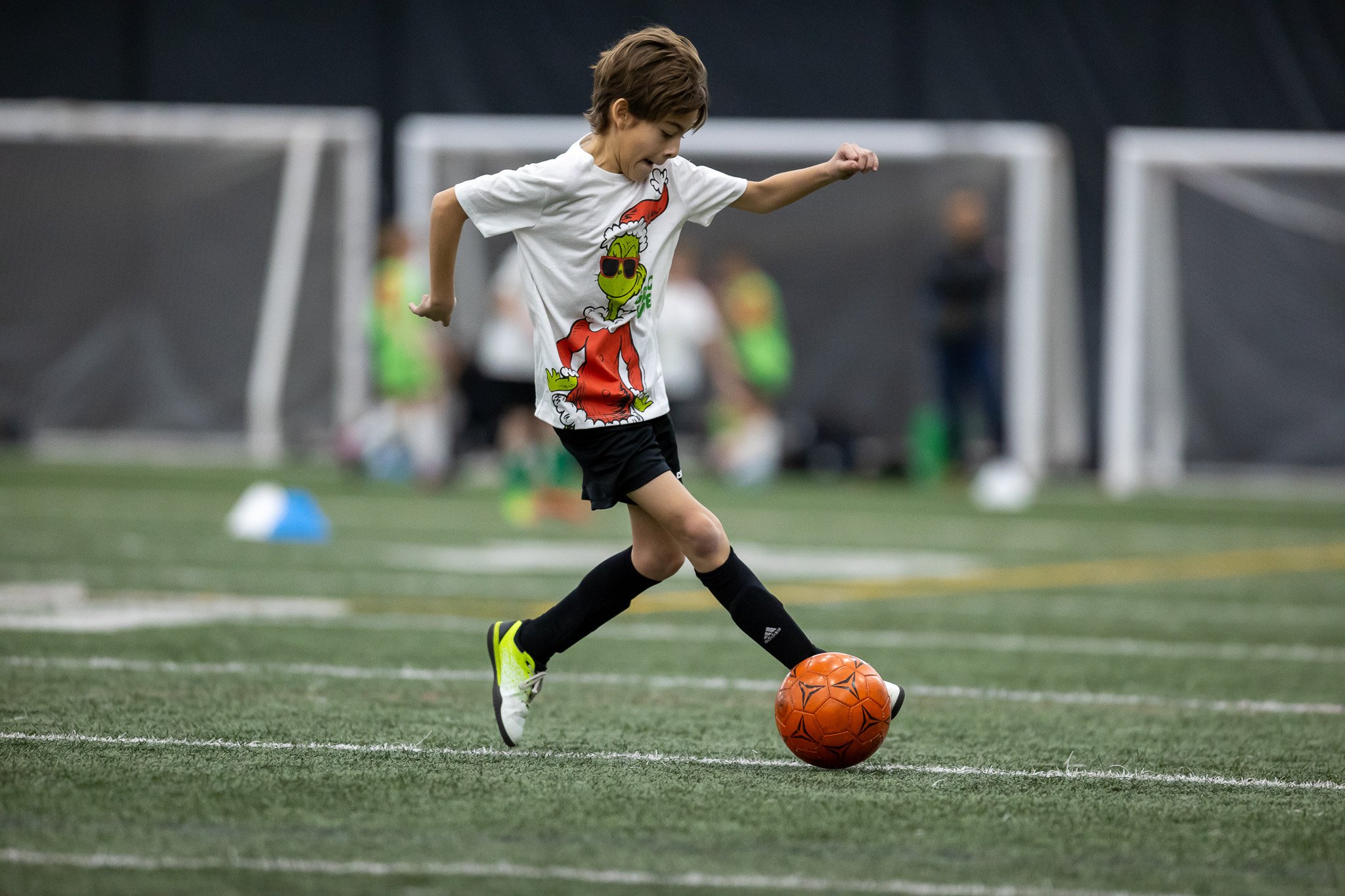 A young boy in a white T-shirt with a cartoon character, black shorts, and bright yellow and black cleats kicks an orange soccer ball on an indoor soccer field.