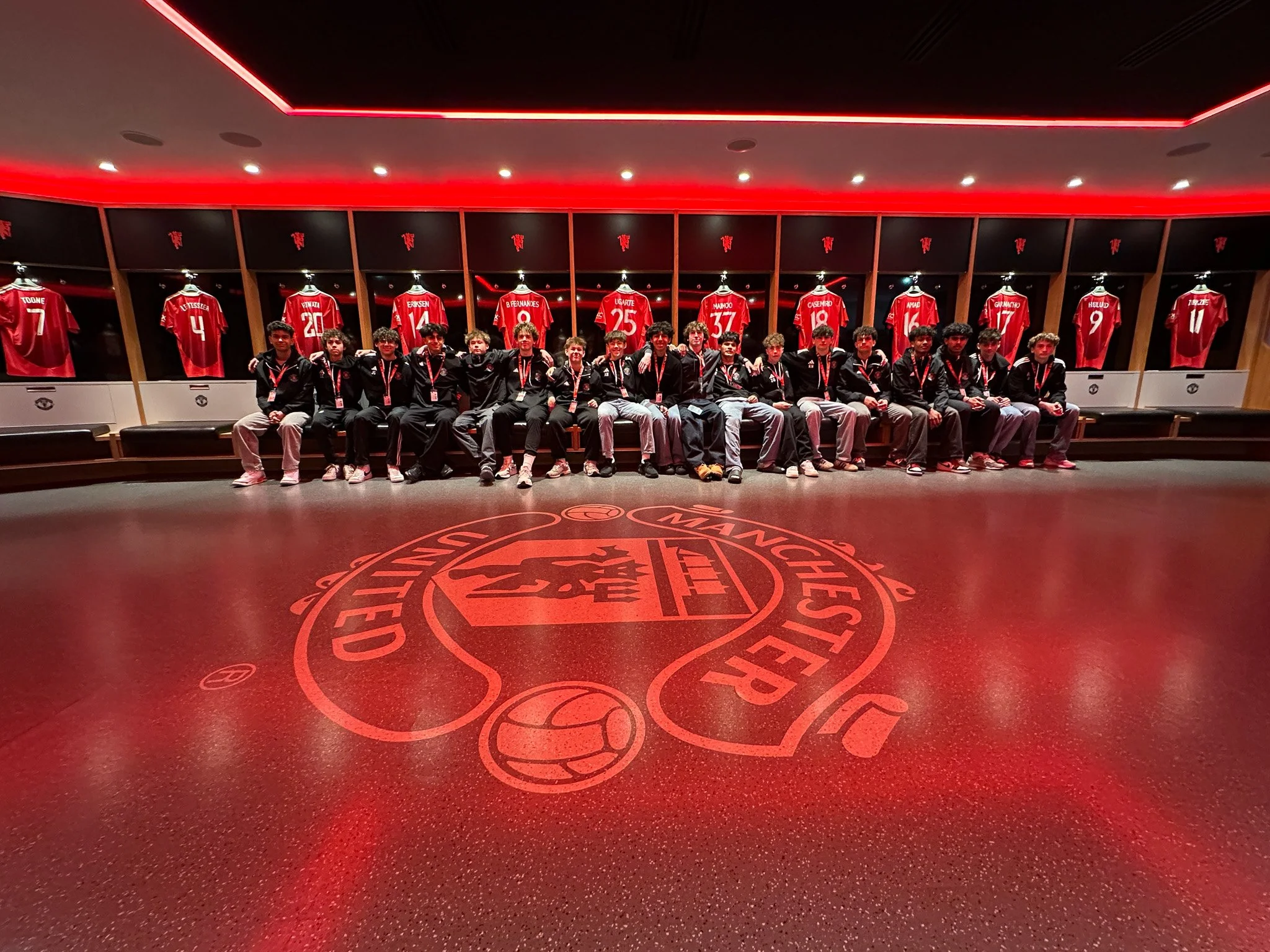 A group of young men sitting on a bench in a locker room with red sports jerseys hanging behind them, featuring the Manchester United logo on the floor.