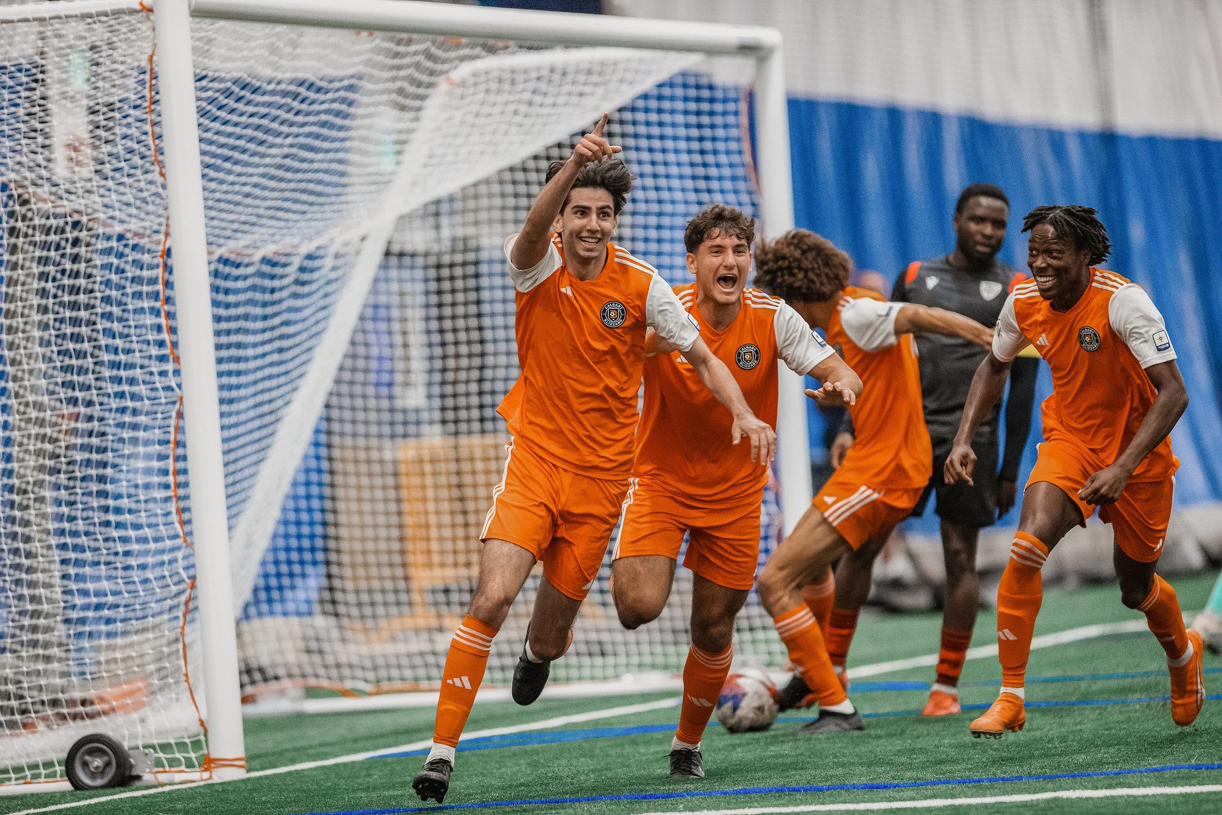 Group of soccer players in orange uniforms celebrating a goal inside an indoor soccer field.