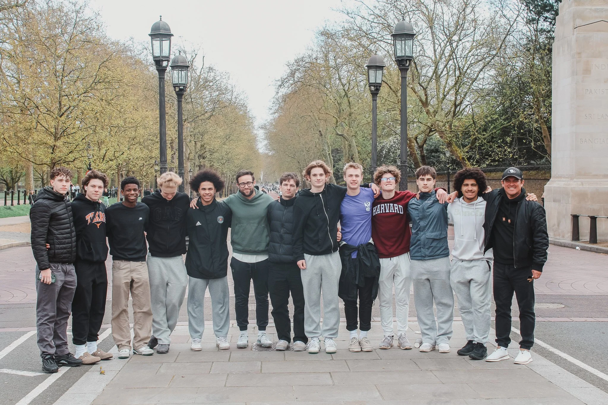 A group of 12 young men standing in a line outdoors on a paved area, with trees and vintage street lamps in the background.