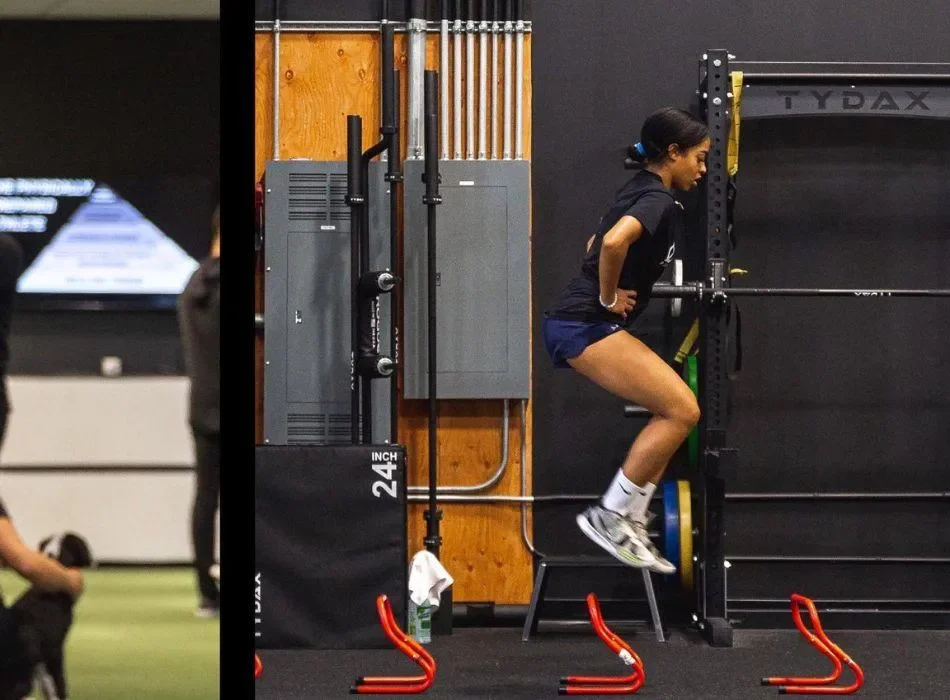 A woman wearing athletic clothing and sneakers is jumping over red hurdles in a gym or training facility.
