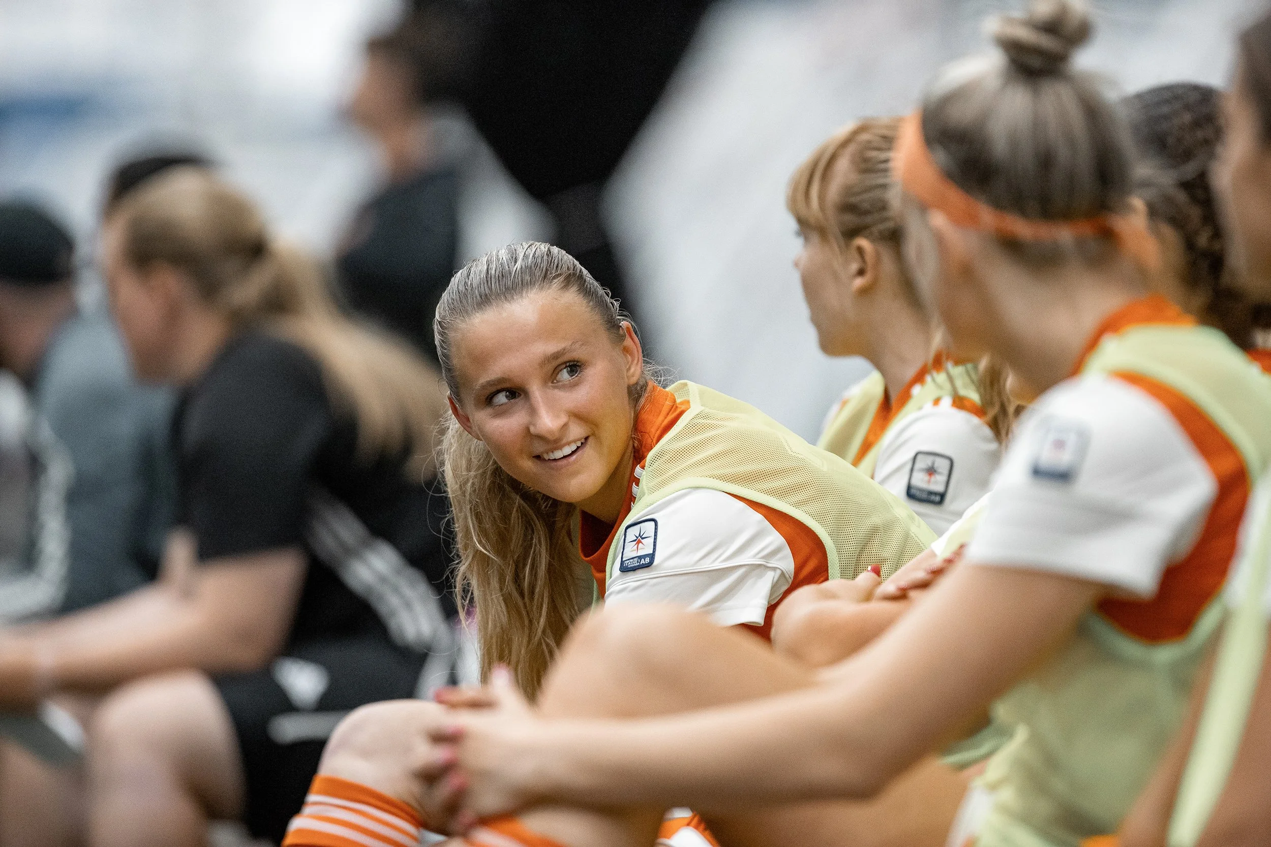 Female soccer players sitting on the bench during a game, wearing yellow and orange jerseys.