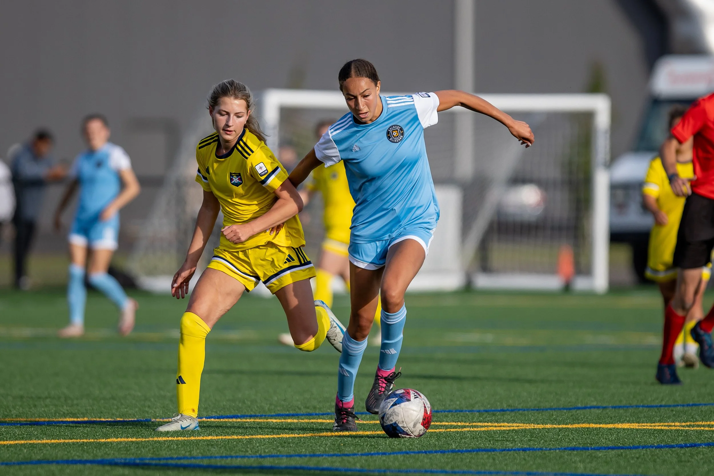 Two women playing soccer on a field, one in a blue uniform and the other in a yellow uniform, competing for the ball.