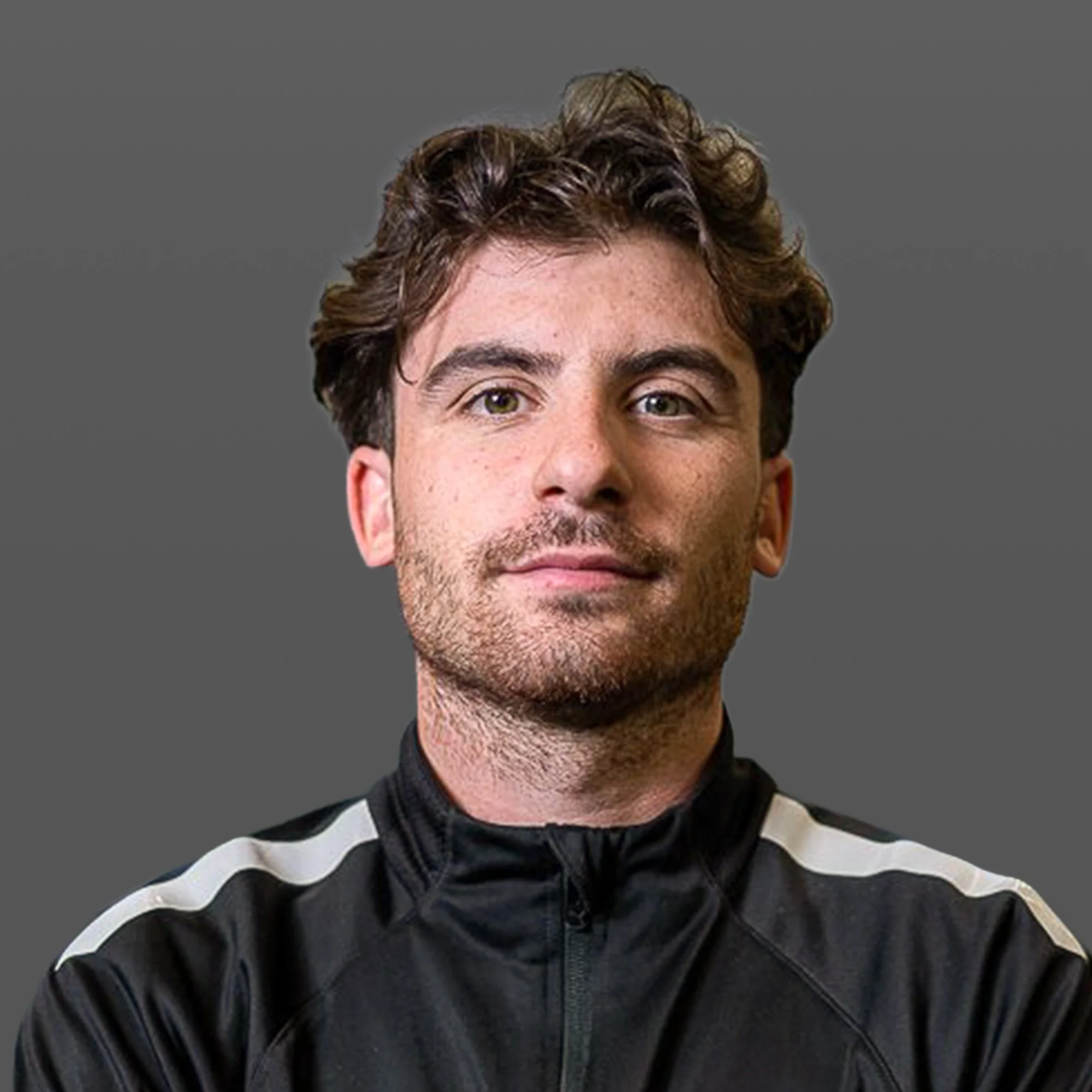 Portrait of a male athlete with brown hair and a beard, wearing a black sports jacket, standing in an indoor sports facility at night with blurred background of a dome-shaped structure and bright lights.