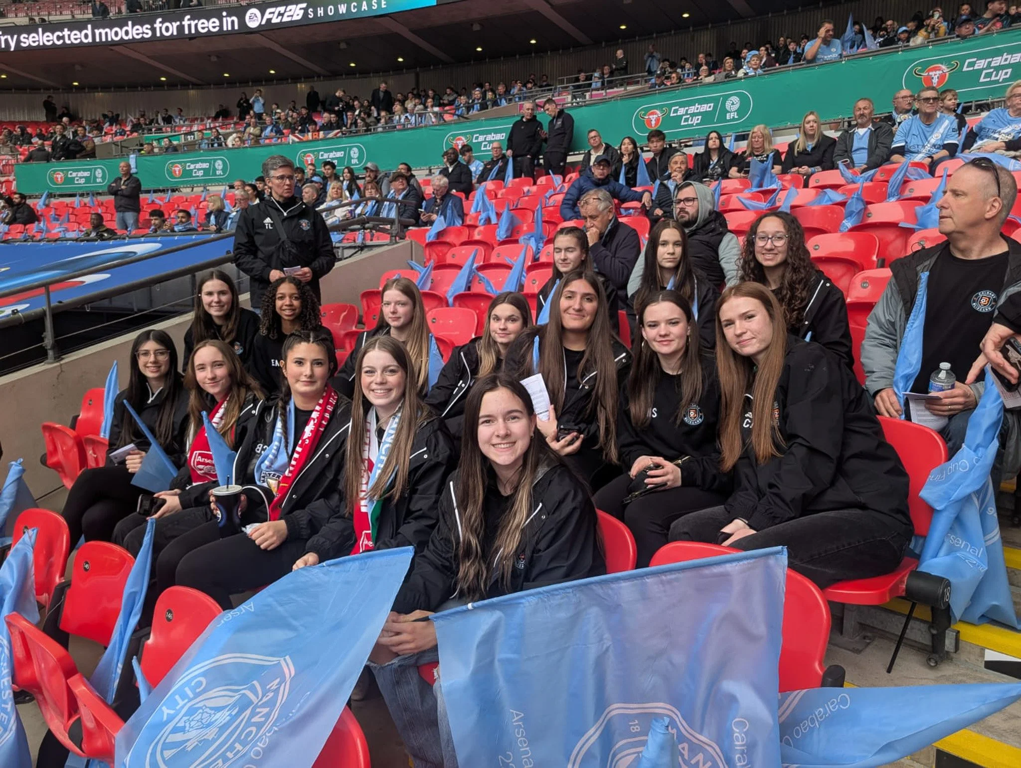 Group of young women sitting in stadium seats with blue flags, dressed in black jackets, at a sporting event with spectators in the background.