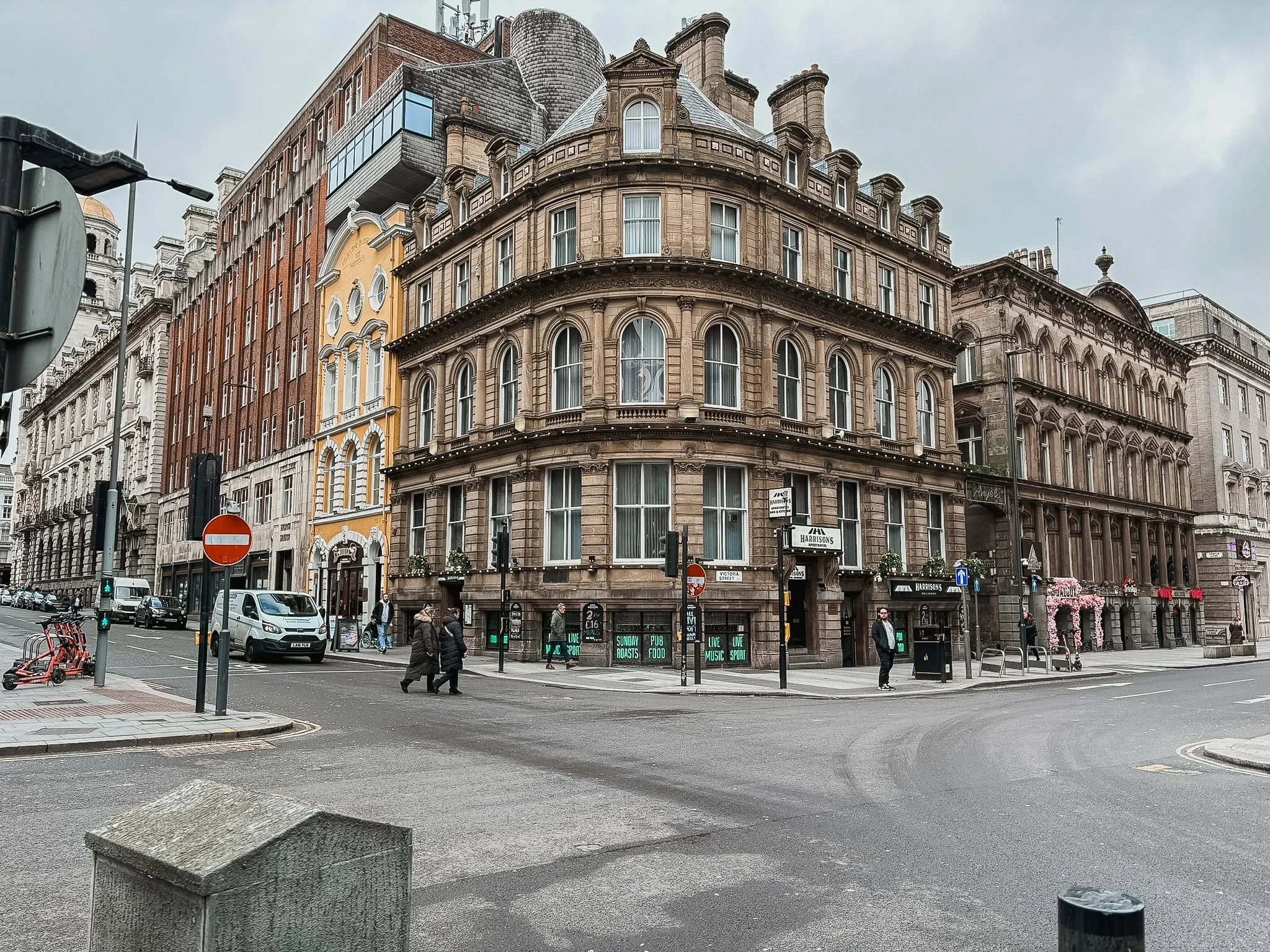A city street corner with historic multi-story buildings, pedestrians, cars, and traffic lights, overcast sky.