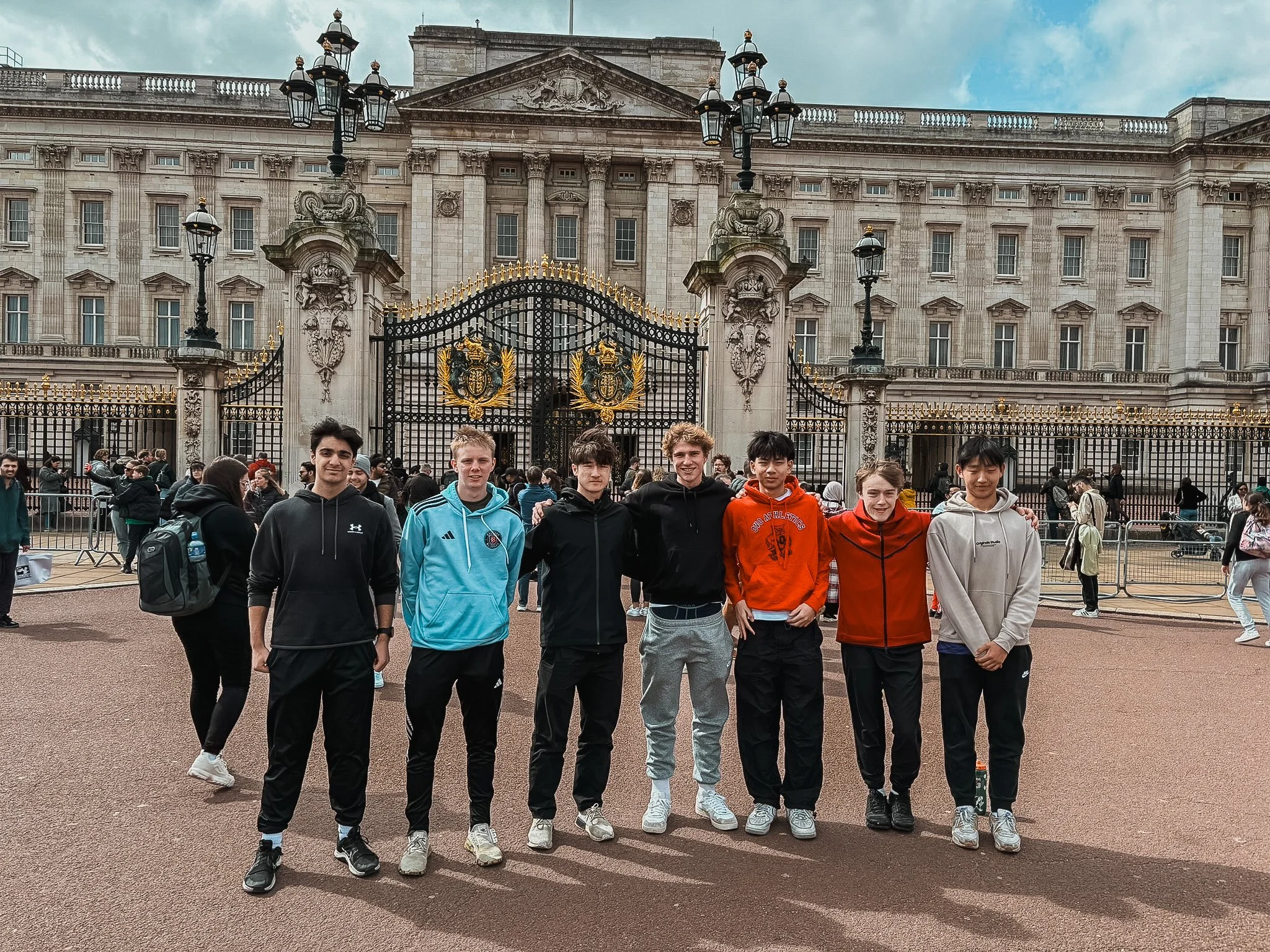 A group of seven young people standing in front of Buckingham Palace with gray overcast sky, some are smiling and wearing casual jackets and pants, with tourists behind a gated entrance to the palace.