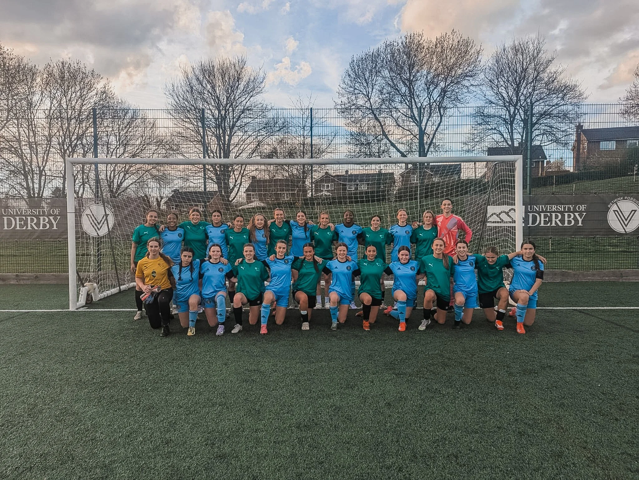 A group of female soccer players, some in turquoise and some in blue jerseys, posing in front of a goal on an outdoor field at the University of Derby. The team is smiling, with some kneeling in the front row and others standing behind. The sky is pa