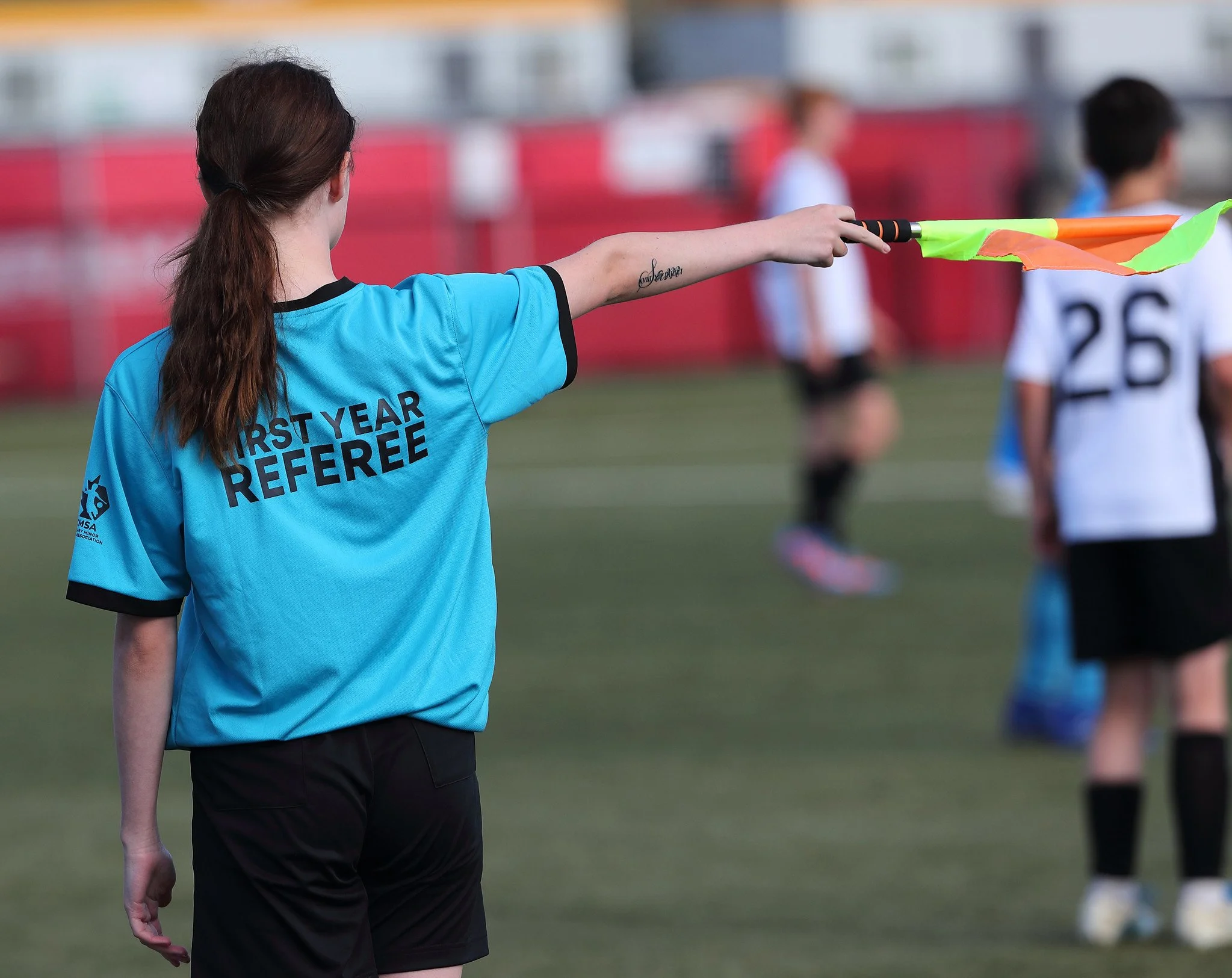 A female first-year soccer referee in a blue shirt and black shorts signals with a flag during a game on a grassy field, with players and red fence in the background.