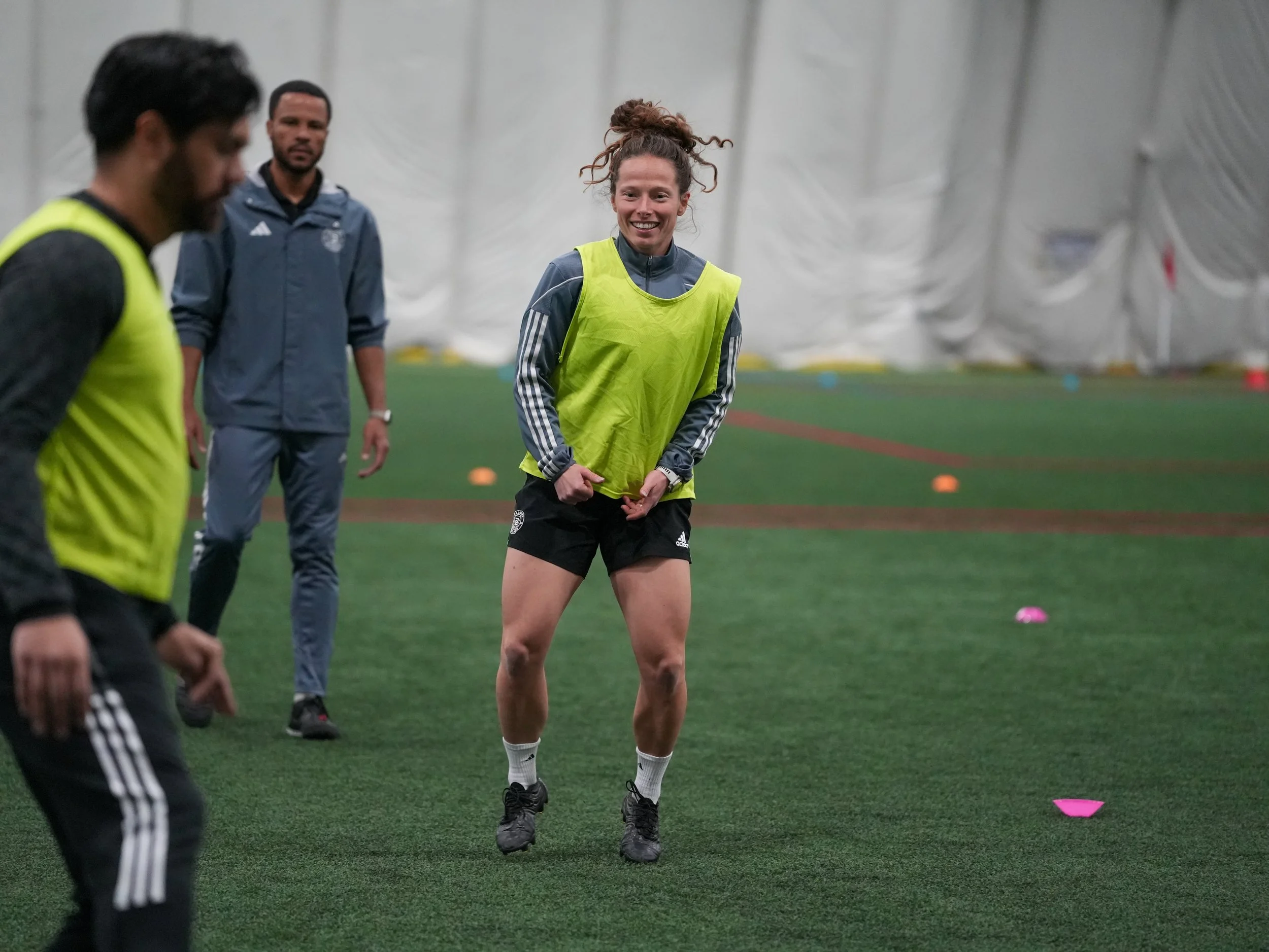Women soccer players training indoors, one smiling woman adjusting her bib, others nearby, orange and pink cones on green turf.