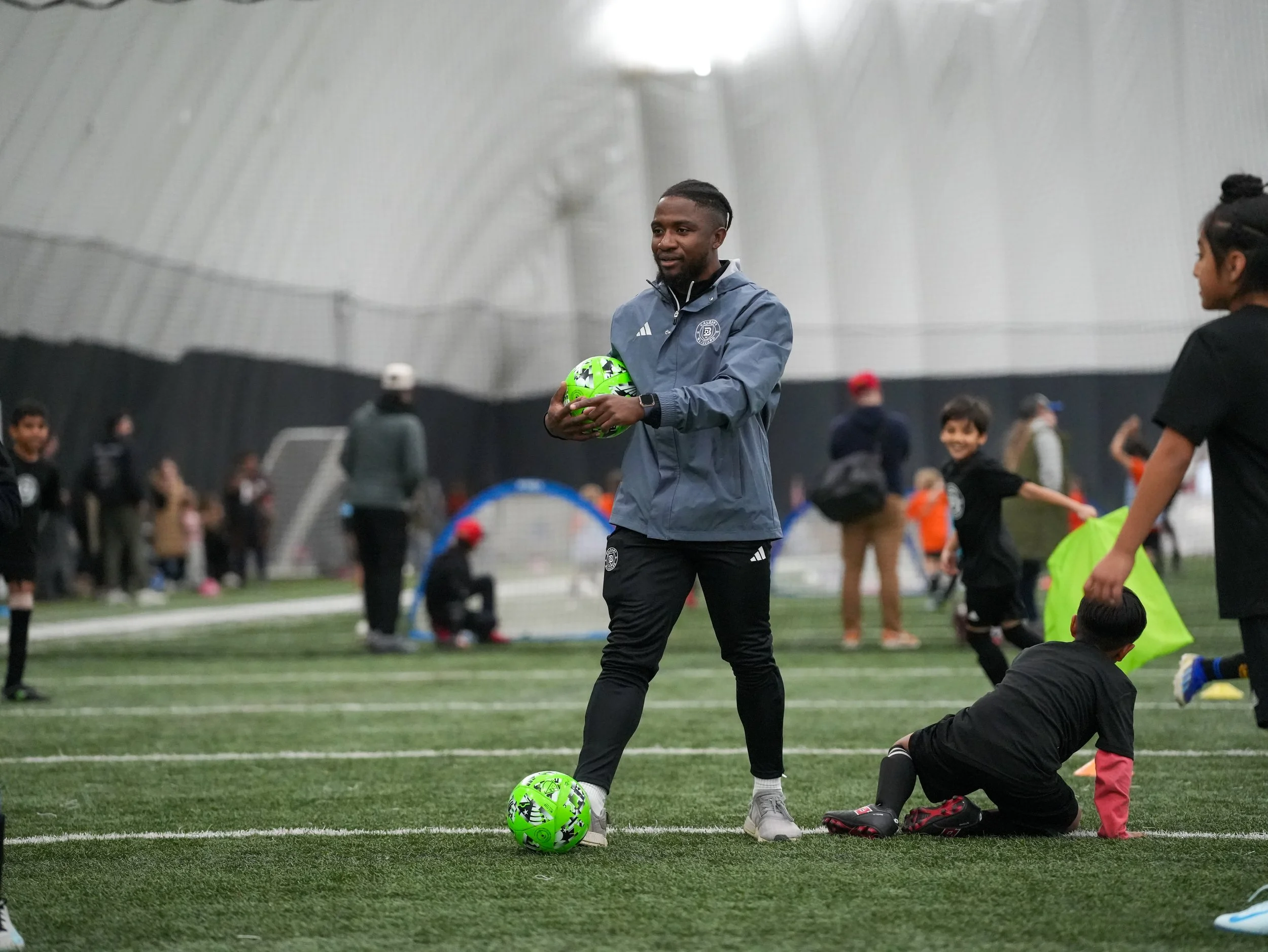 A soccer coach, wearing a gray jacket, stands on an indoor soccer field holding a green and black soccer ball. Children, some with soccer gear, are playing and running in the background inside a large domed sports facility.
