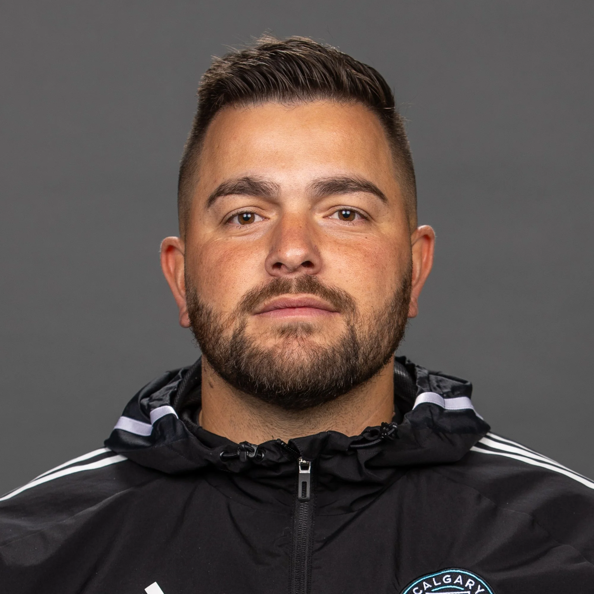 A man with short hair and a beard wearing a black Adidas jacket with a police badge on the chest, standing outdoors at night.