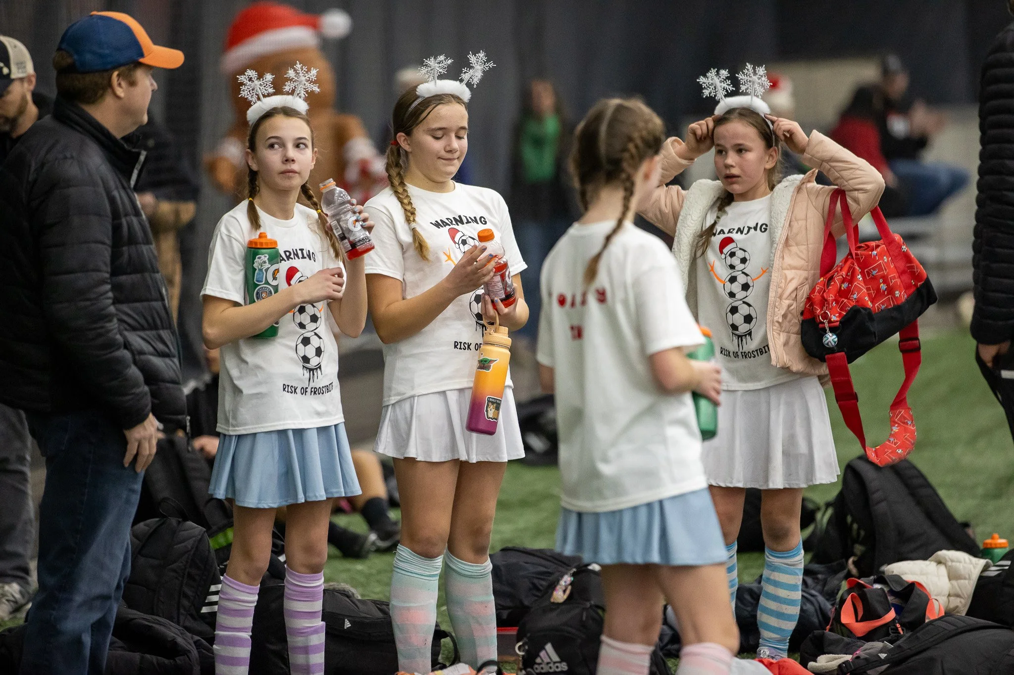 Group of young girls in soccer uniforms and Christmas-themed headbands standing indoors, some holding water bottles, with adults and backpacks in the background.