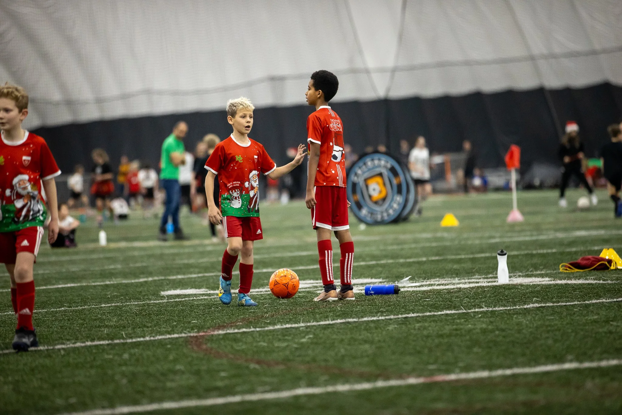 Two young boys in red soccer uniforms talking on an indoor soccer field, one of them setting down an orange soccer ball, with other children and adults in the background.